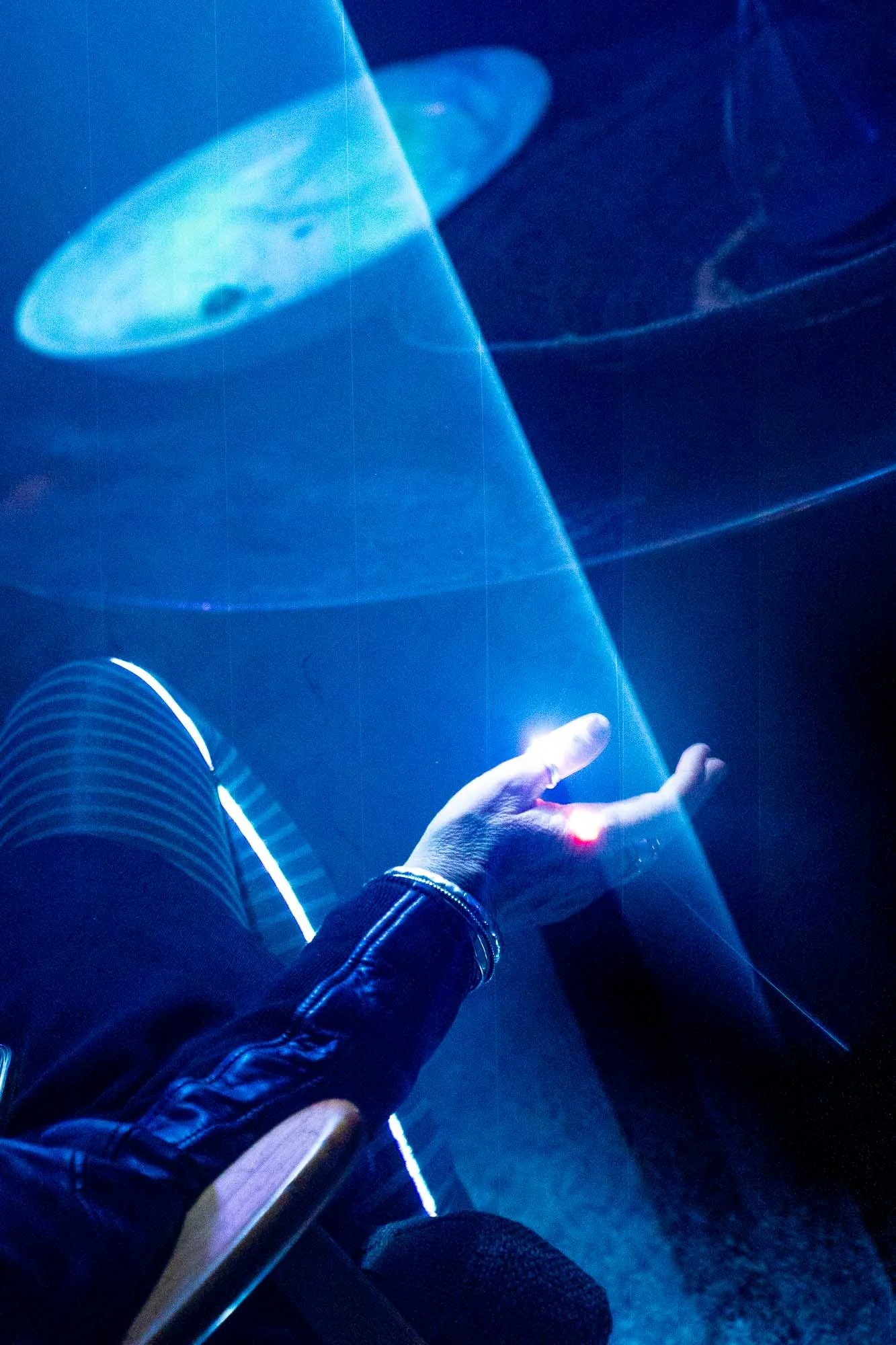 Person wearing a leather jacket and striped pants seated in a dark room illuminated by blue LED lights, with a finger extended towards the light.
