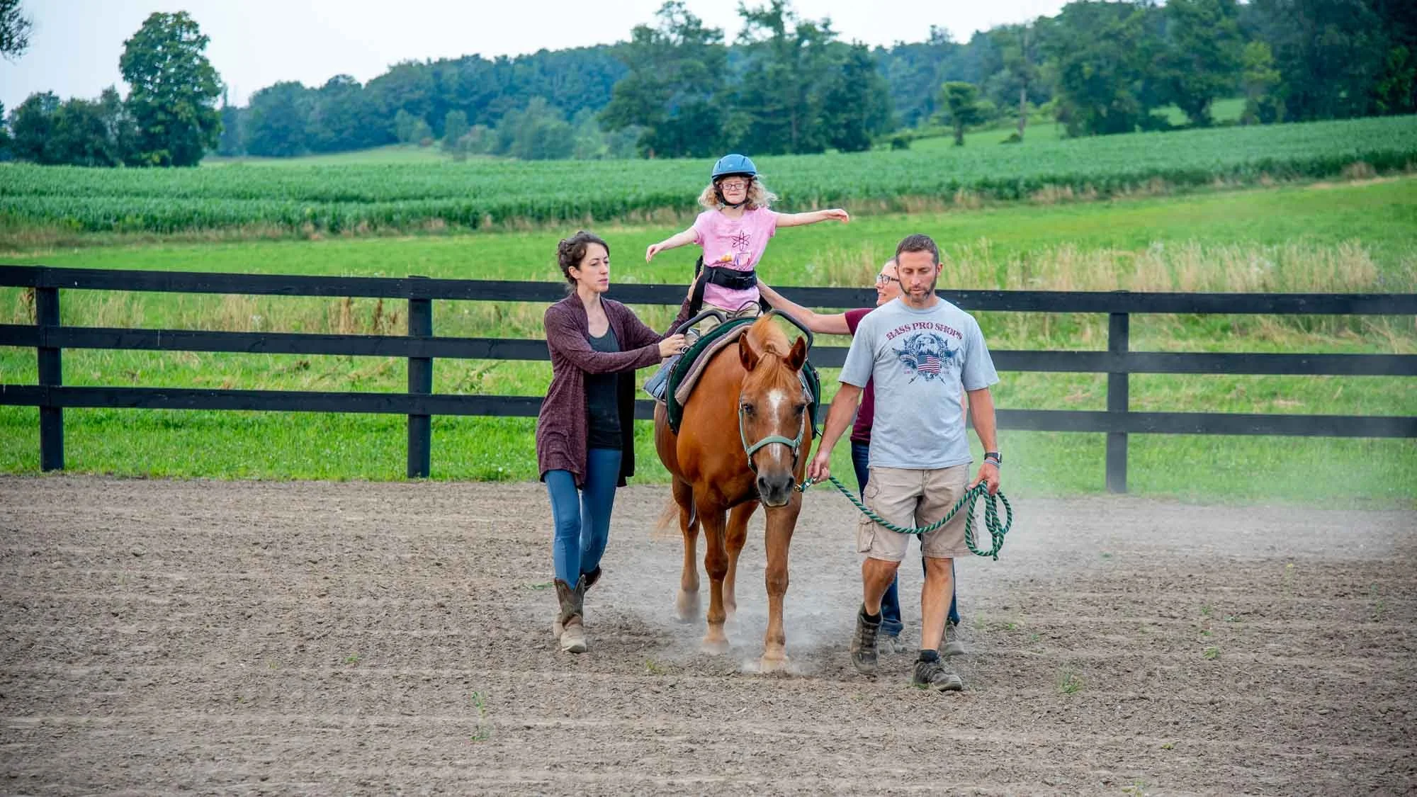 Four people with a small child on horseback at an outdoor riding area with green fields and trees in the background.