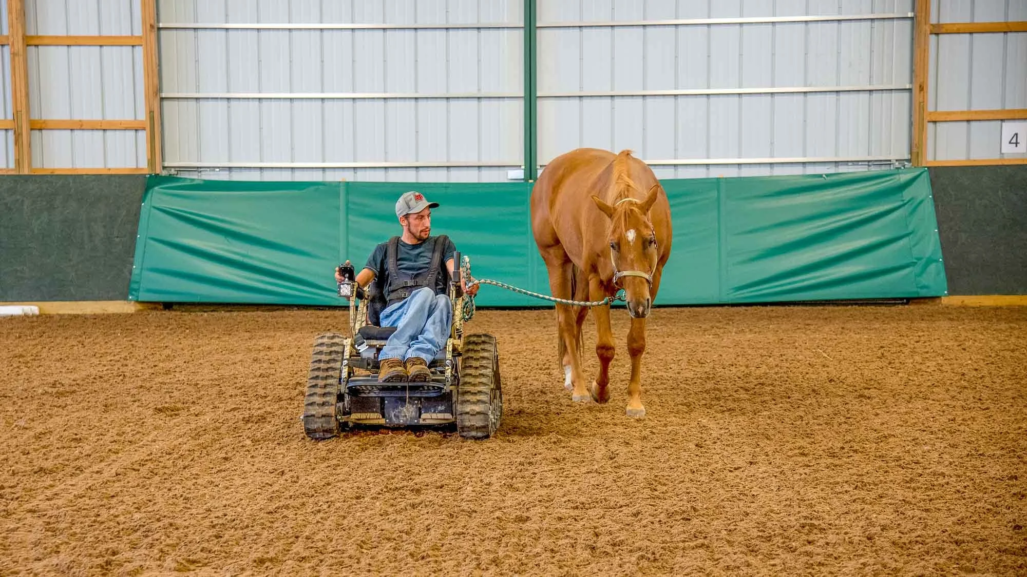A man in a wheelchair leading a chestnut horse with a white blaze in an indoor riding arena.