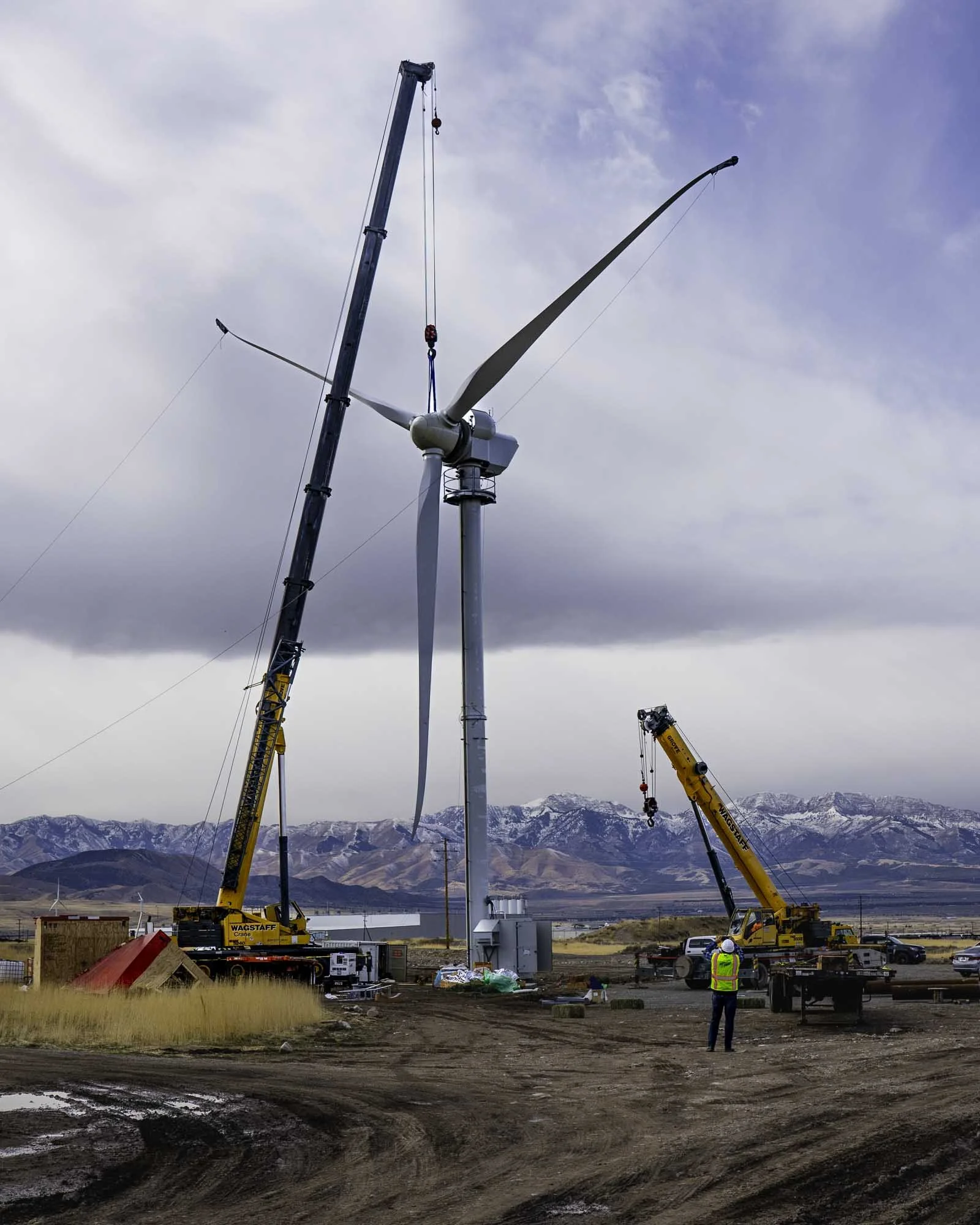 Construction workers install a large wind turbine in a mountainous area with a cloudy sky. Two cranes lift parts of the turbine, and a person in a safety vest observes the work.