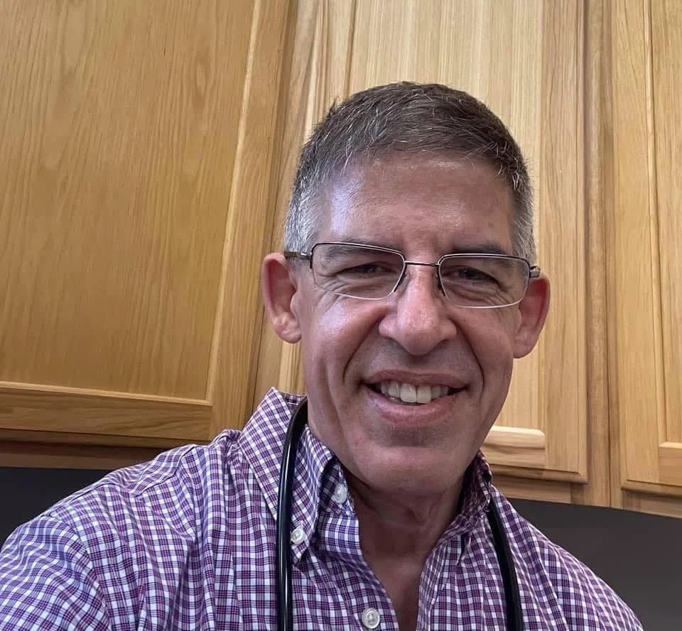 A smiling man wearing glasses and a purple checkered shirt with a stethoscope around his neck, standing in front of wooden cabinets.