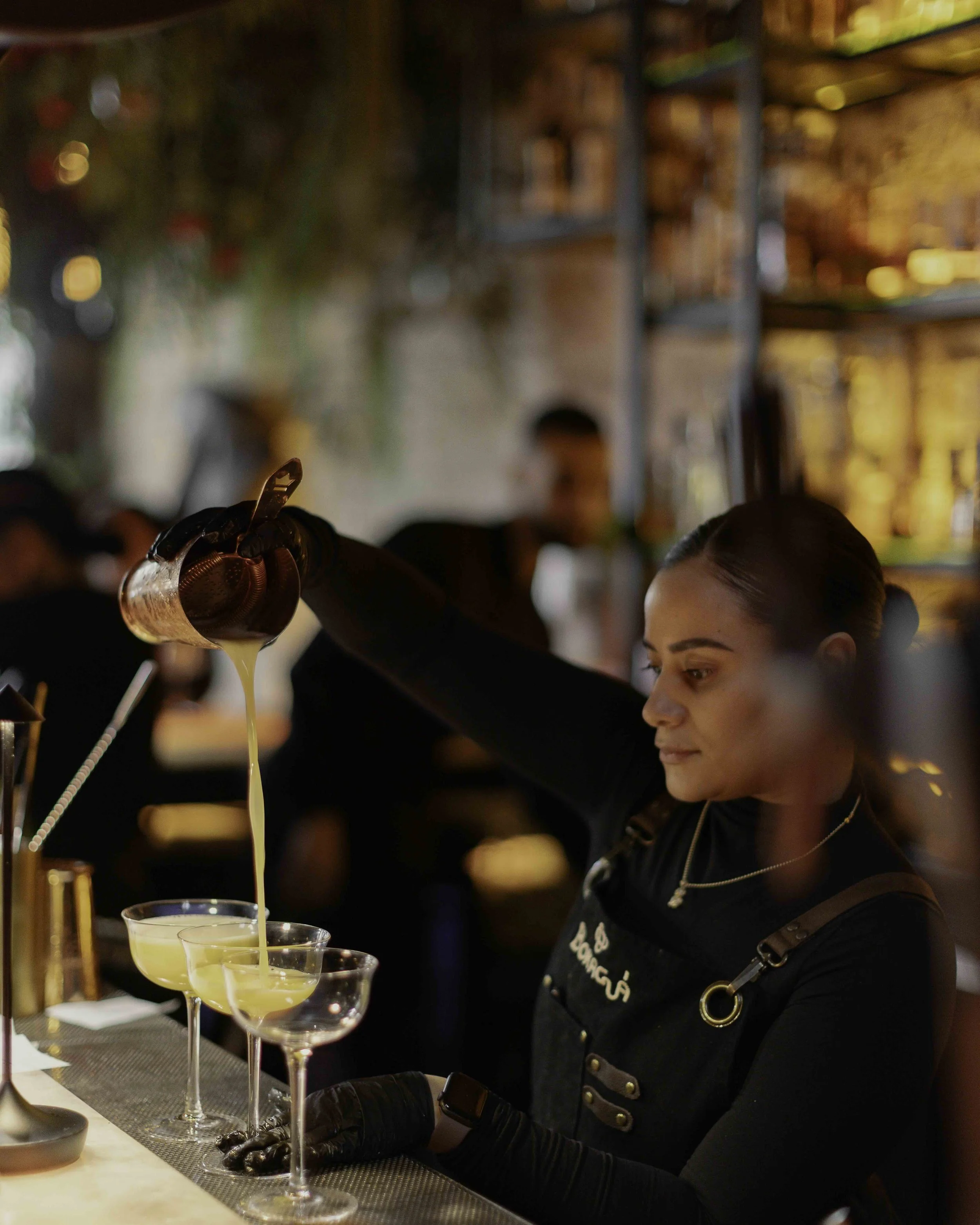 A female bartender pouring a yellow cocktail into champagne glasses in a bar with warm lighting and blurred background.