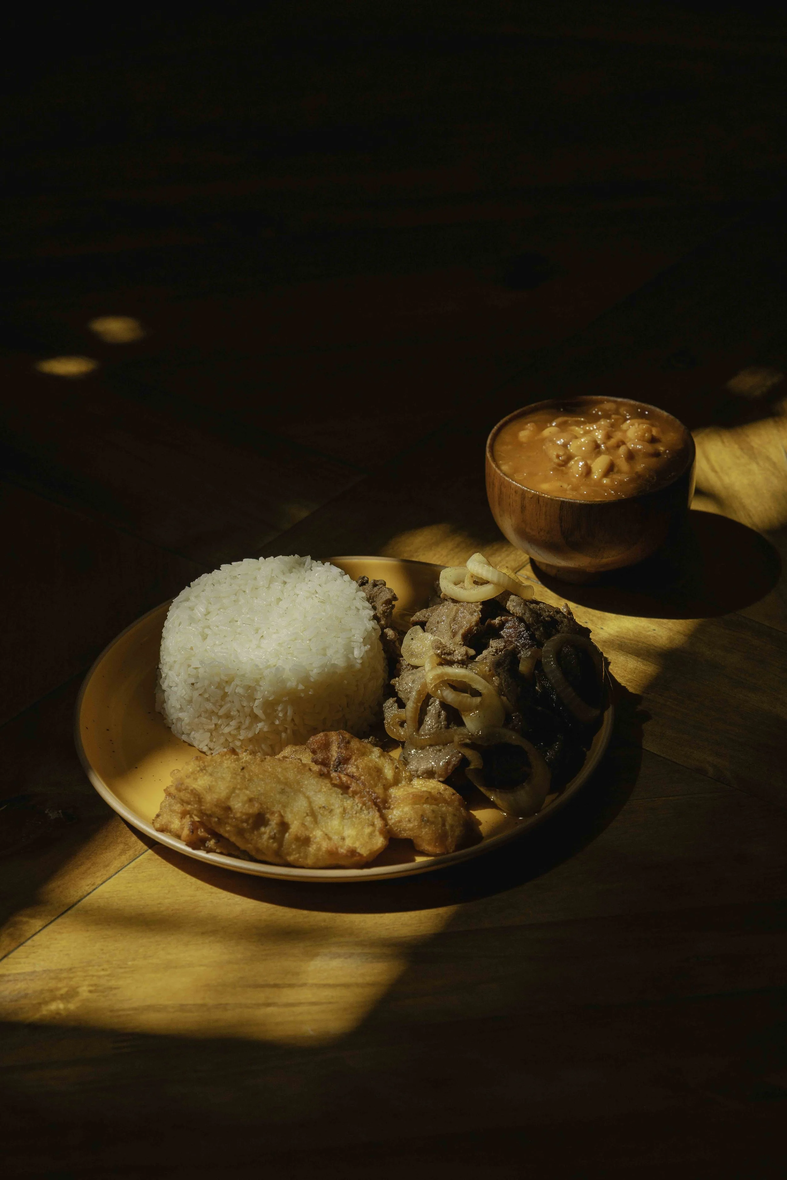 Plate of white rice, fried chicken, beef with onions, and a bowl of bean soup on a wooden table with shadows.