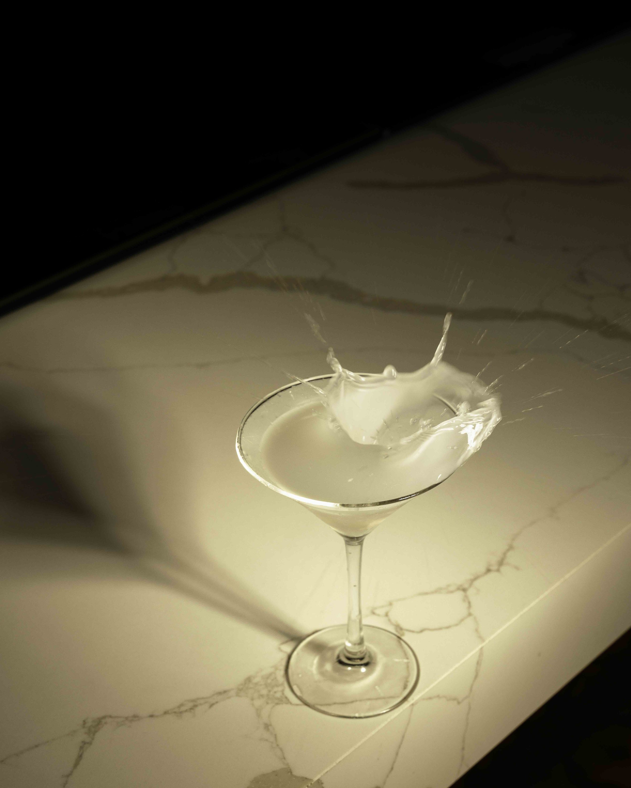 A martini glass with a splash of liquid, captured mid-motion, on a white marble surface with a dark background.