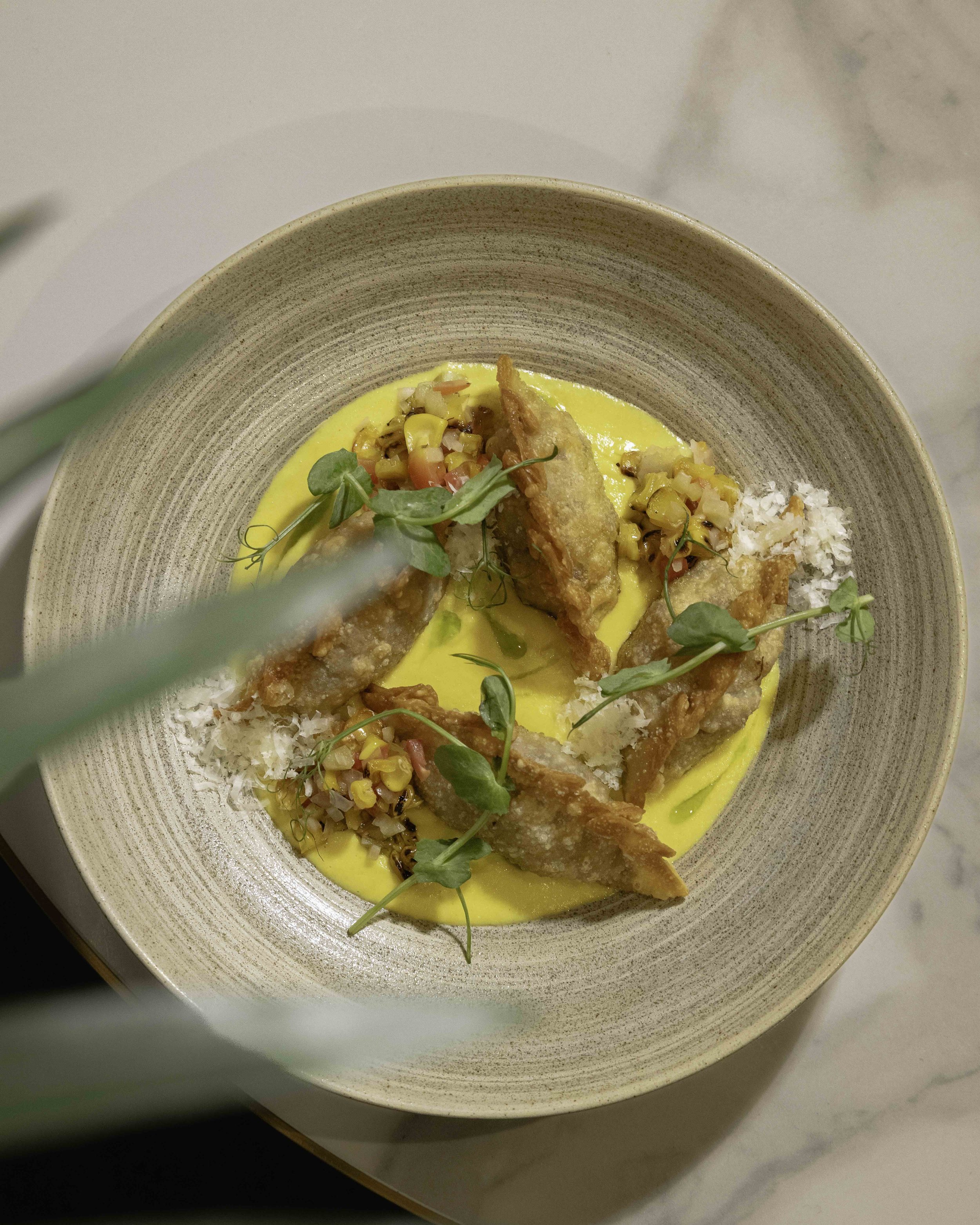 A bowl of food with fried fish fillets, microgreens, a yellow sauce, and a garnish of chopped vegetables in a decorative bowl.