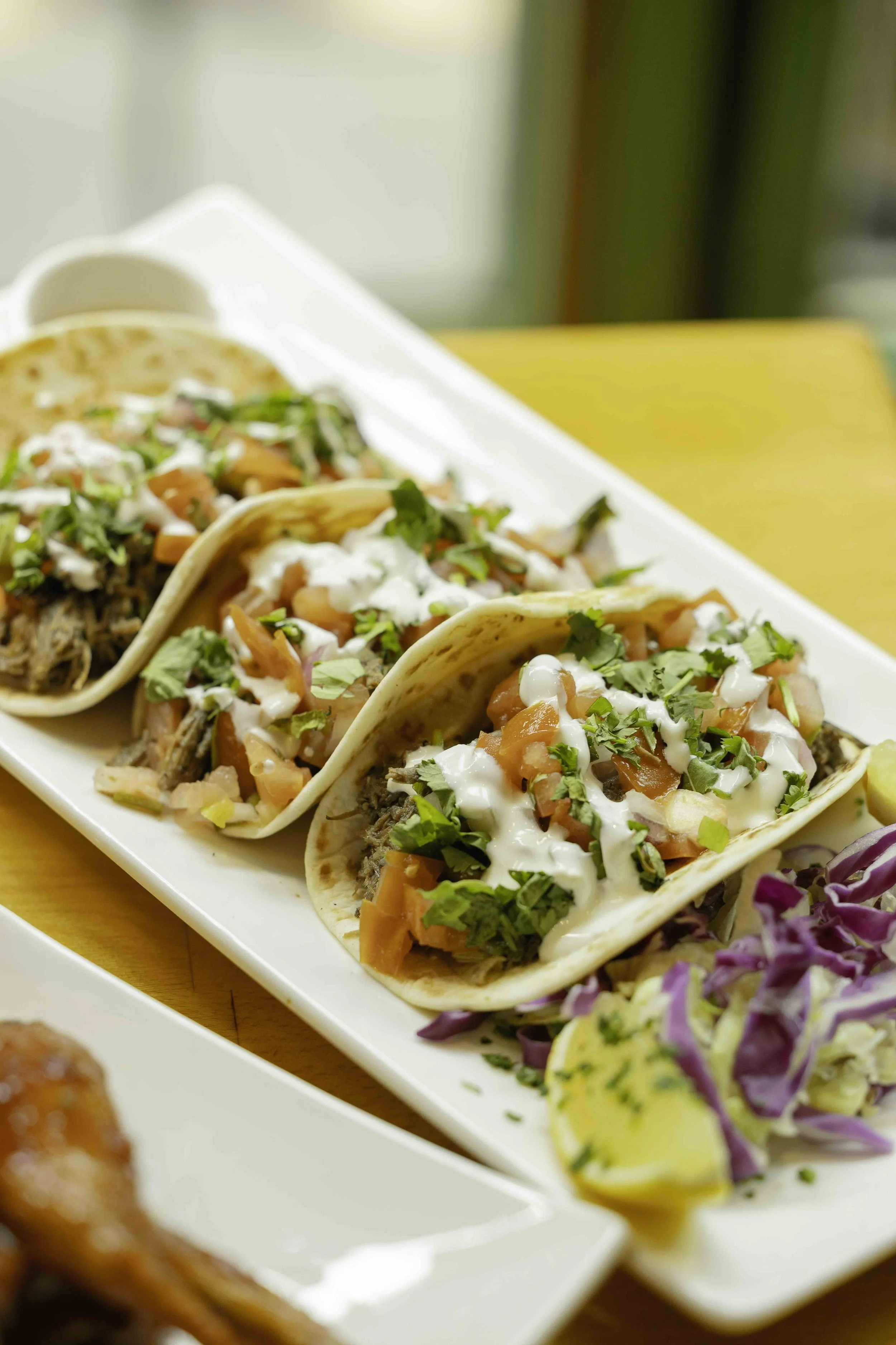 Close-up of three tacos filled with shredded meat, topped with chopped vegetables, herbs, and white sauce, arranged on a white serving platter.