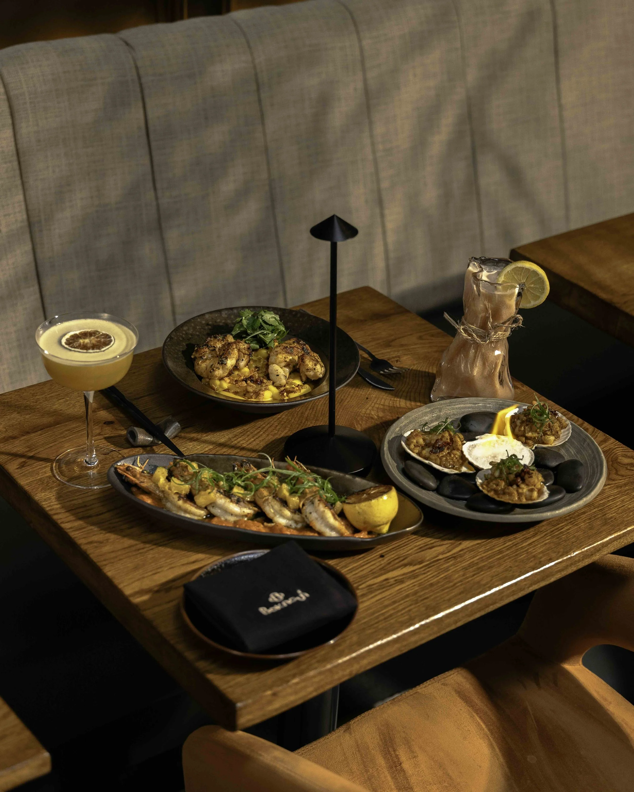 A wooden table set with various gourmet dishes, a cocktail with a lemon garnish, a glass of yellow beverage with a citrus garnish, and a pink drink with a lemon slice, arranged in an upscale restaurant environment.