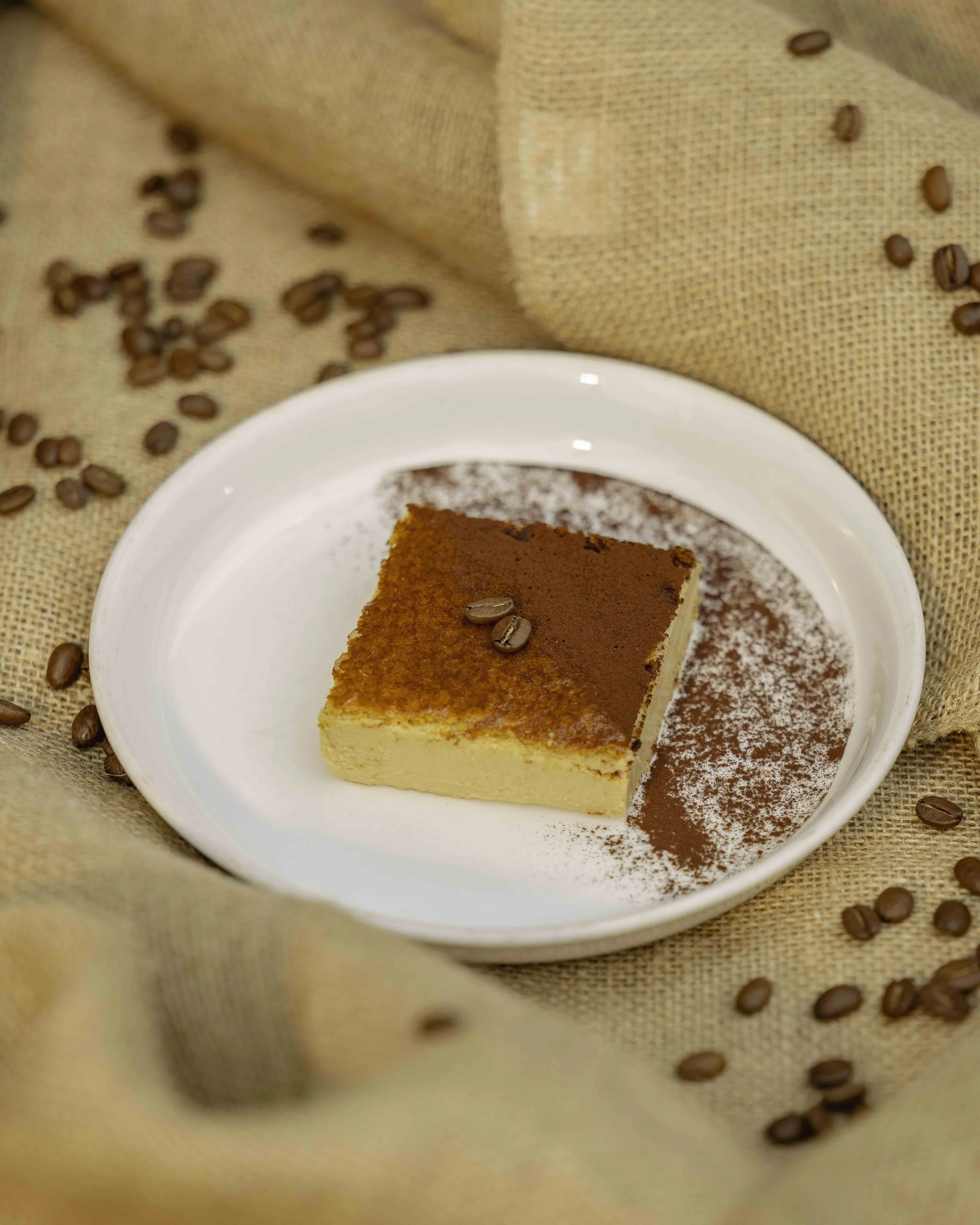 A slice of cheesecake on a white plate decorated with cocoa powder and two coffee beans, with more coffee beans scattered on a beige fabric background.
