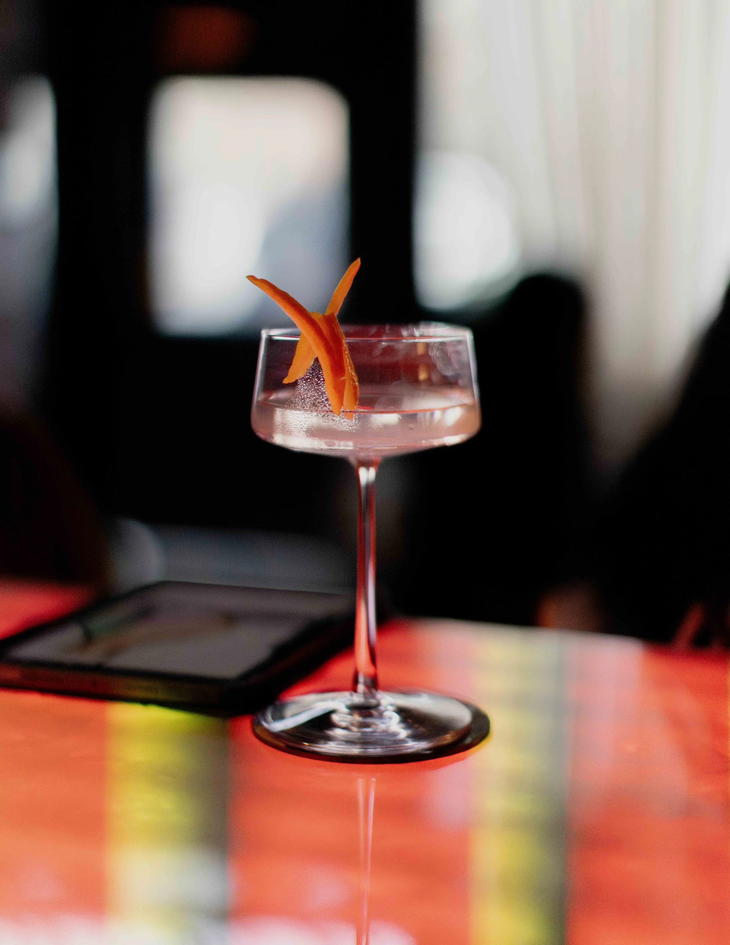A pink cocktail with orange twists garnish in a coupe glass on a reflective table with blurred background.