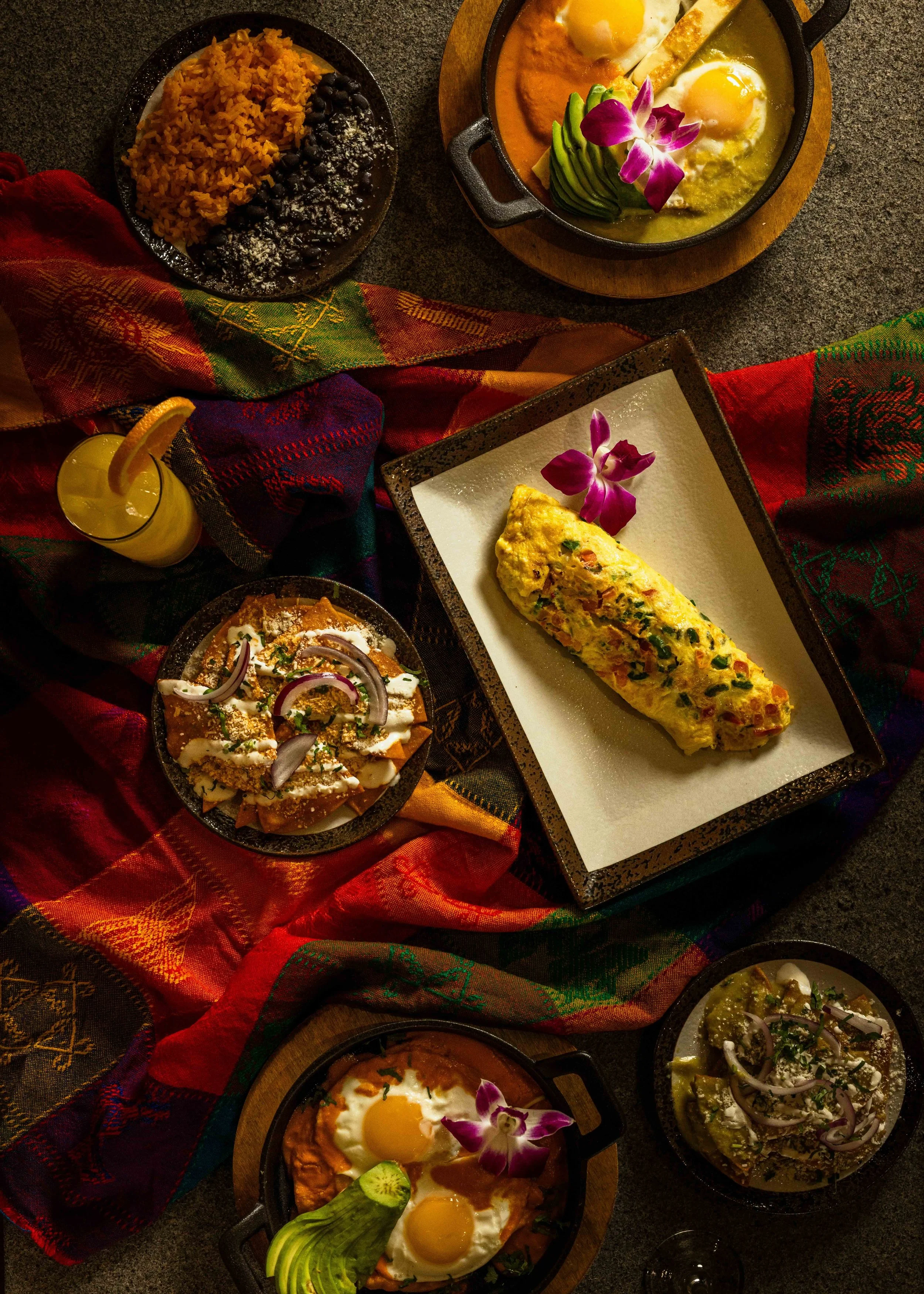 A table with various Mexican dishes including a rice and black beans bowl, a frittata with vegetables, two eggs with sauce, guacamole with lime and onion, and two bowls of salad, all on a colorful cloth.