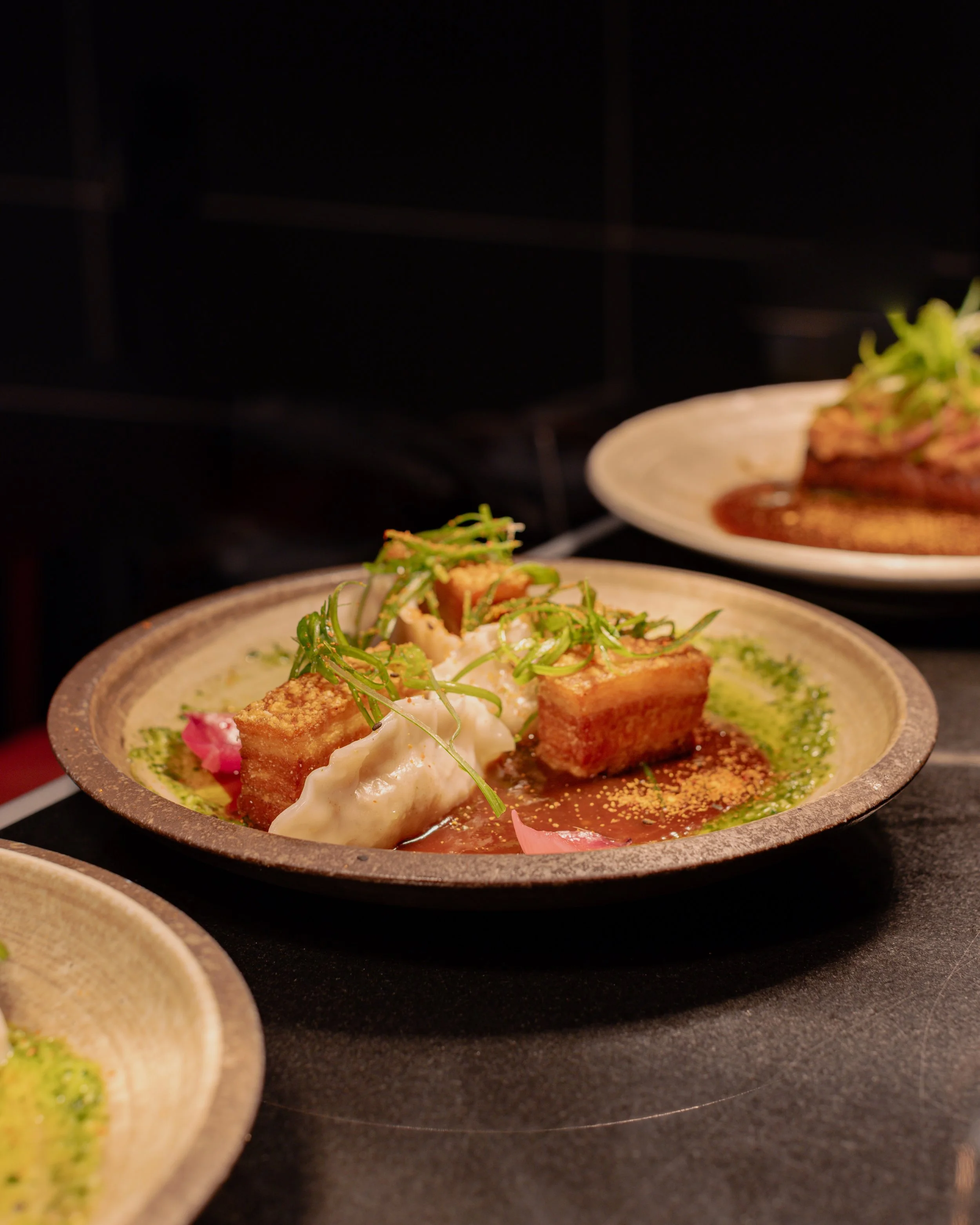 A plated gourmet dish with fried tofu, green herbs, and sauce on a dark surface.