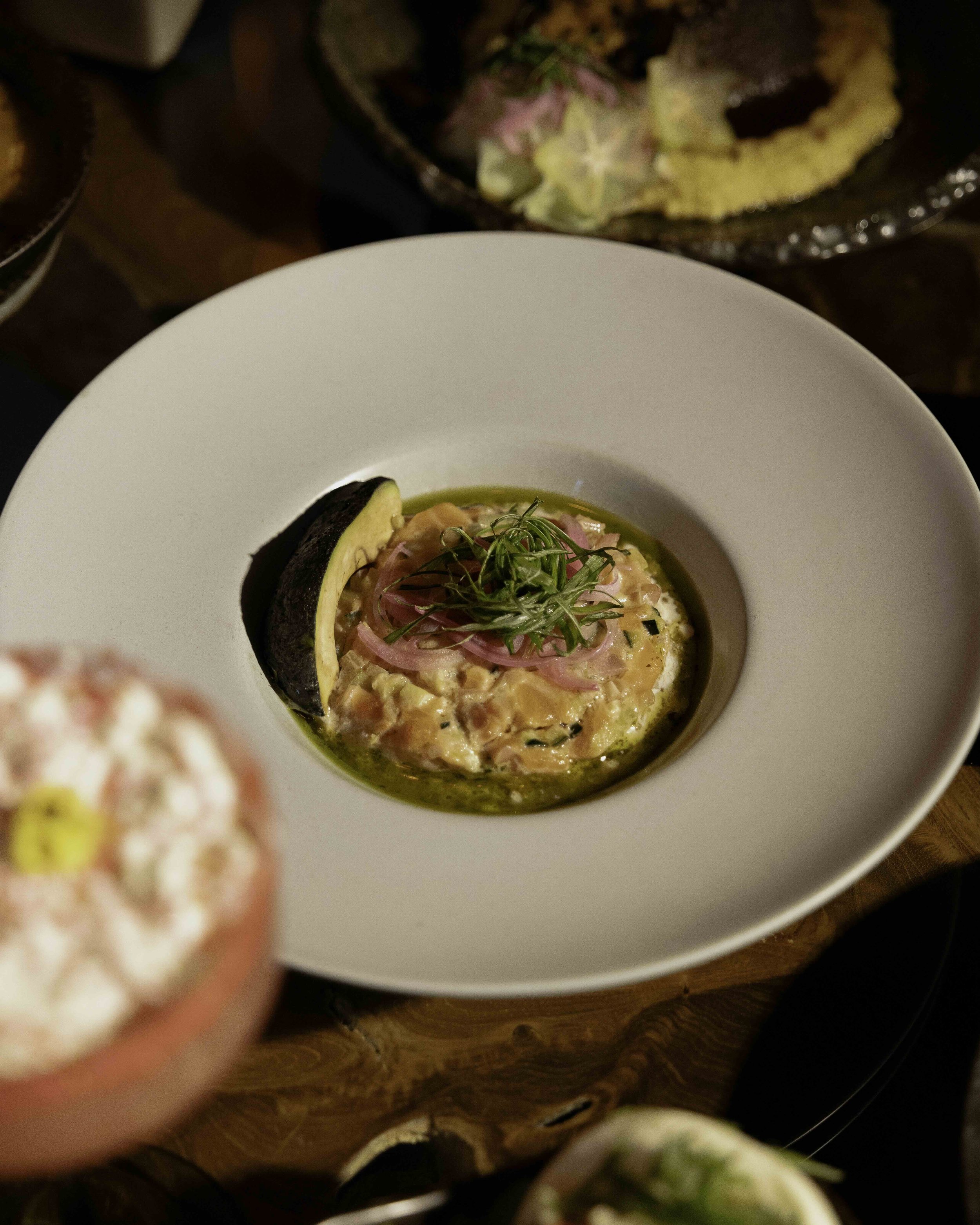 A white bowl containing a dish of raw salmon mixed with herbs, topped with pickled red onions and microgreens, garnished with a lemon wedge and olive oil.