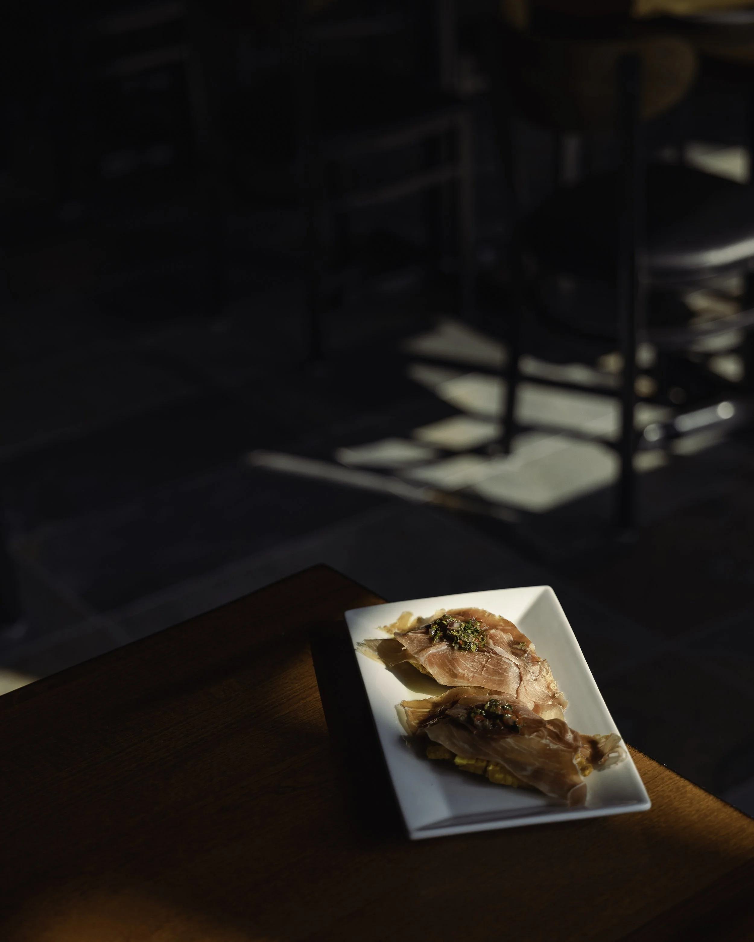 A white rectangular plate with three slices of prosciutto garnished with herbs, resting on a wooden table in a dimly lit setting.