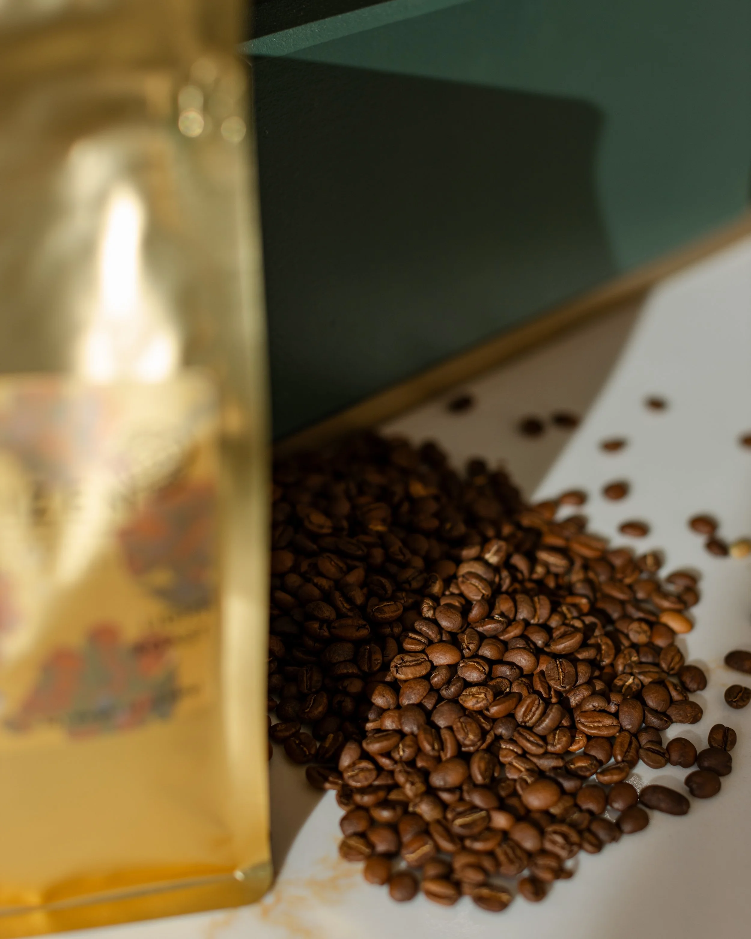 A pile of roasted coffee beans on a white surface with some scattered beans nearby, partially obscured by a gold-colored bag of coffee.
