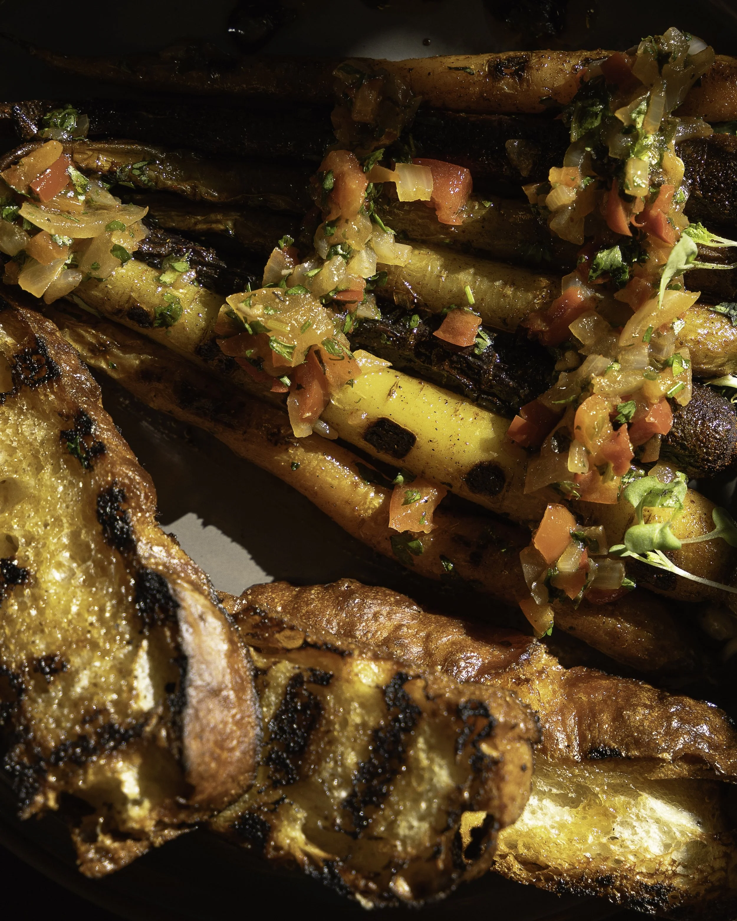 Close-up of grilled vegetables, including carrots and potatoes, topped with diced tomatoes, onions, and fresh herbs, with a piece of grilled bread in the foreground.