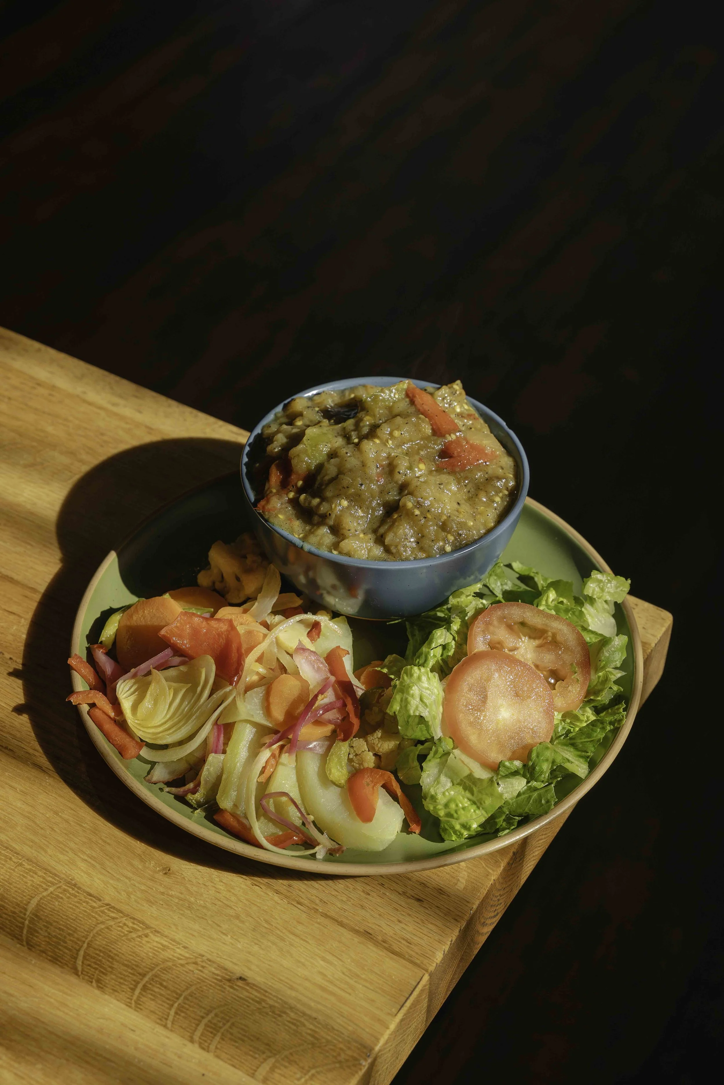 A plate with a variety of fresh vegetables including sliced tomatoes, lettuce, sliced onions, and shredded carrots, with a bowl of stew nearby, set on a wooden surface.