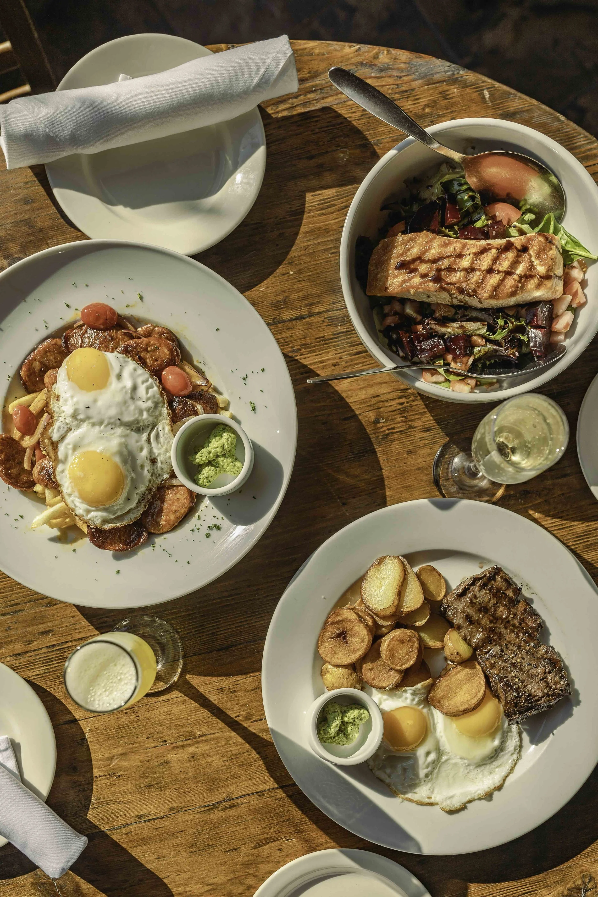 Overhead view of a wooden table with two plates of breakfast: one with eggs, sausage, and potatoes, and another with steak, fried eggs, and potatoes. A bowl with salad and grilled salmon, a glass of sparkling wine, and a small glass with a yellow dri