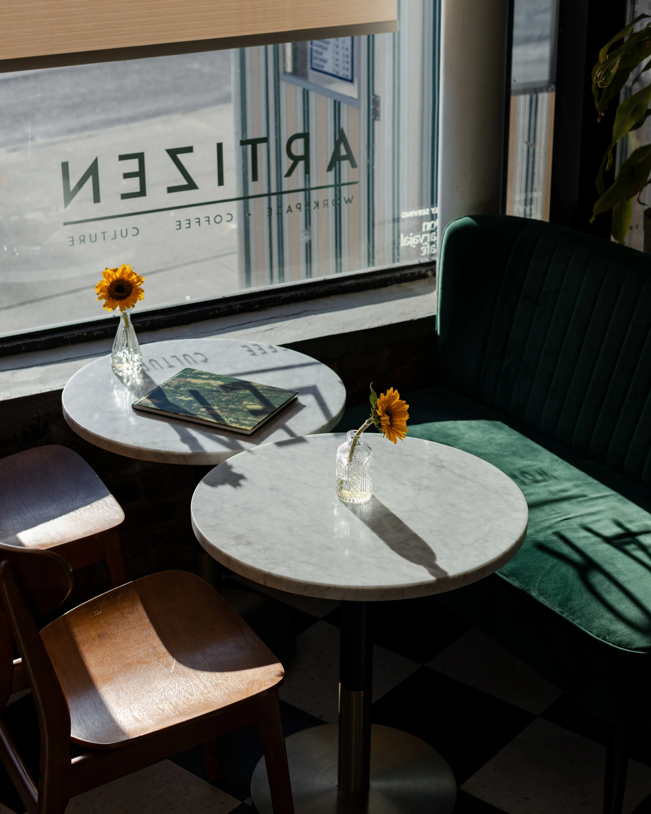 Indoor seating with two white marble tables, sunflower vases, a book, a wooden chair, a green cushioned bench, sunlight streaming through a window, and a sign with the word 'ARTIZEN' on the window.