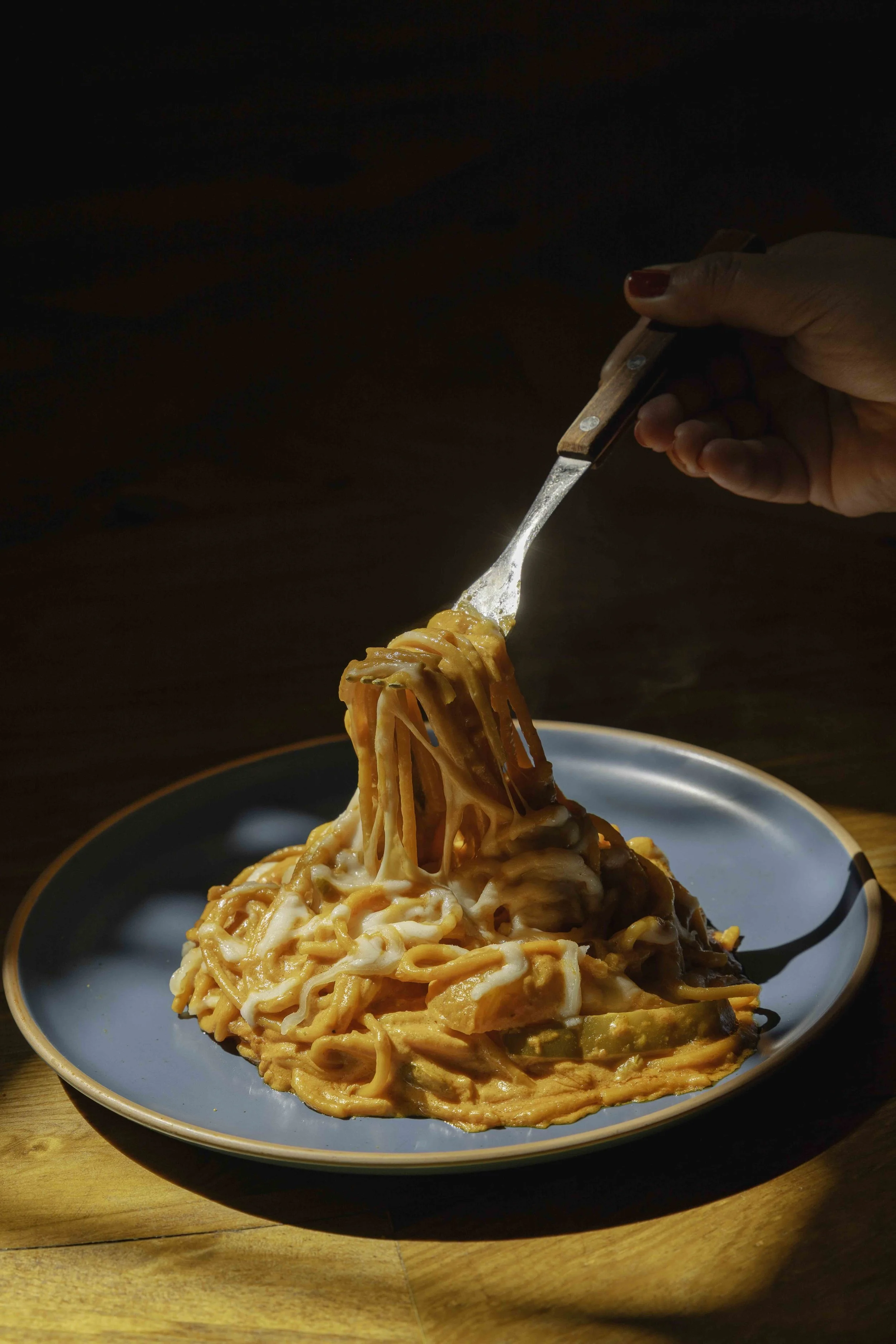 A plate of cheesy spaghetti with vegetables, with a hand lifting a forkful of pasta.