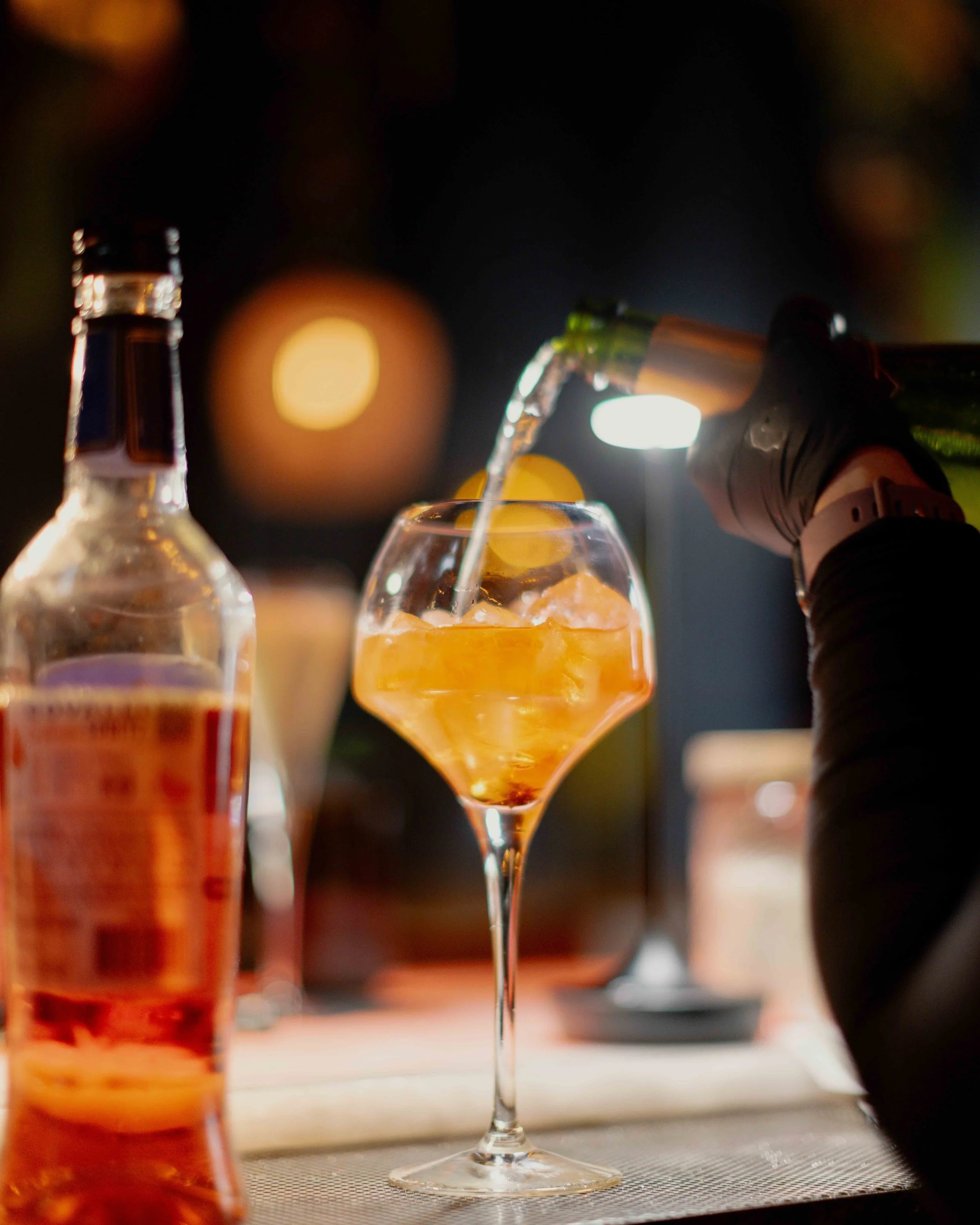 Person pouring a drink into a cocktail glass with ice at a bar or restaurant, with bottles and warm lighting in the background.