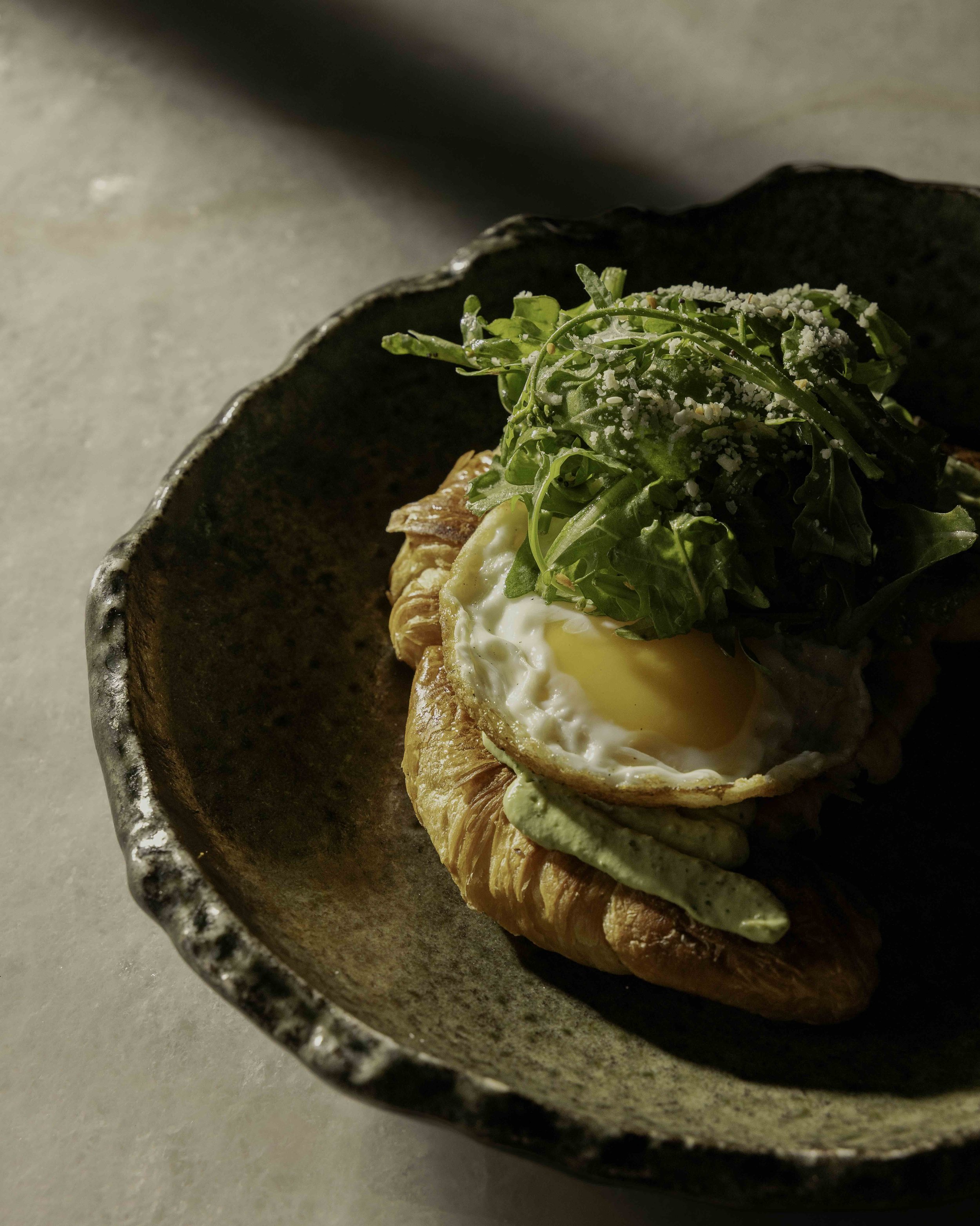 Croissant topped with a fried egg, avocado spread, leafy greens, and grated cheese in a dark ceramic bowl.