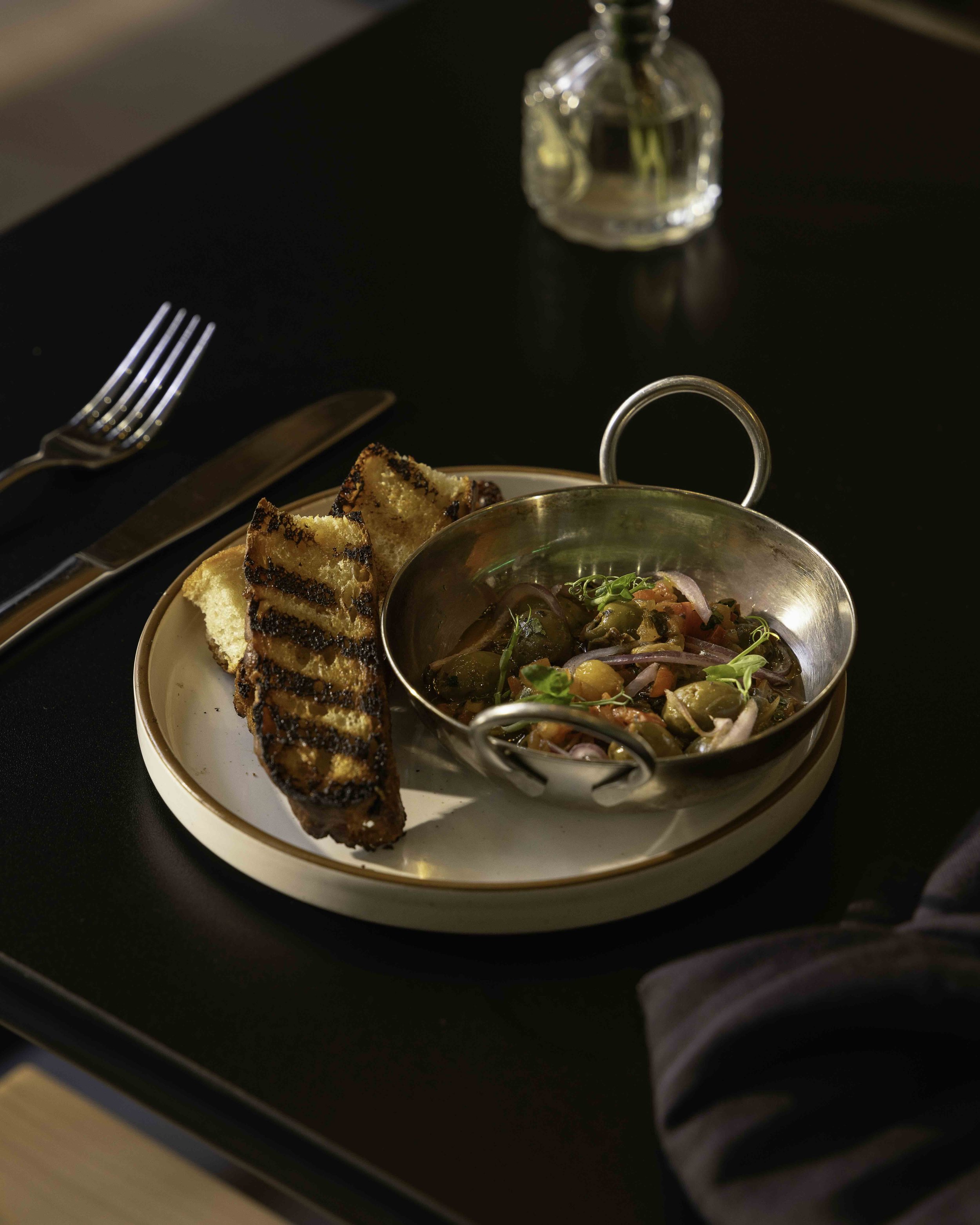 Grilled bread and vegetable salad served in a metal bowl on a white plate, placed on a black table with a fork, knife, and a small glass vase in the background.