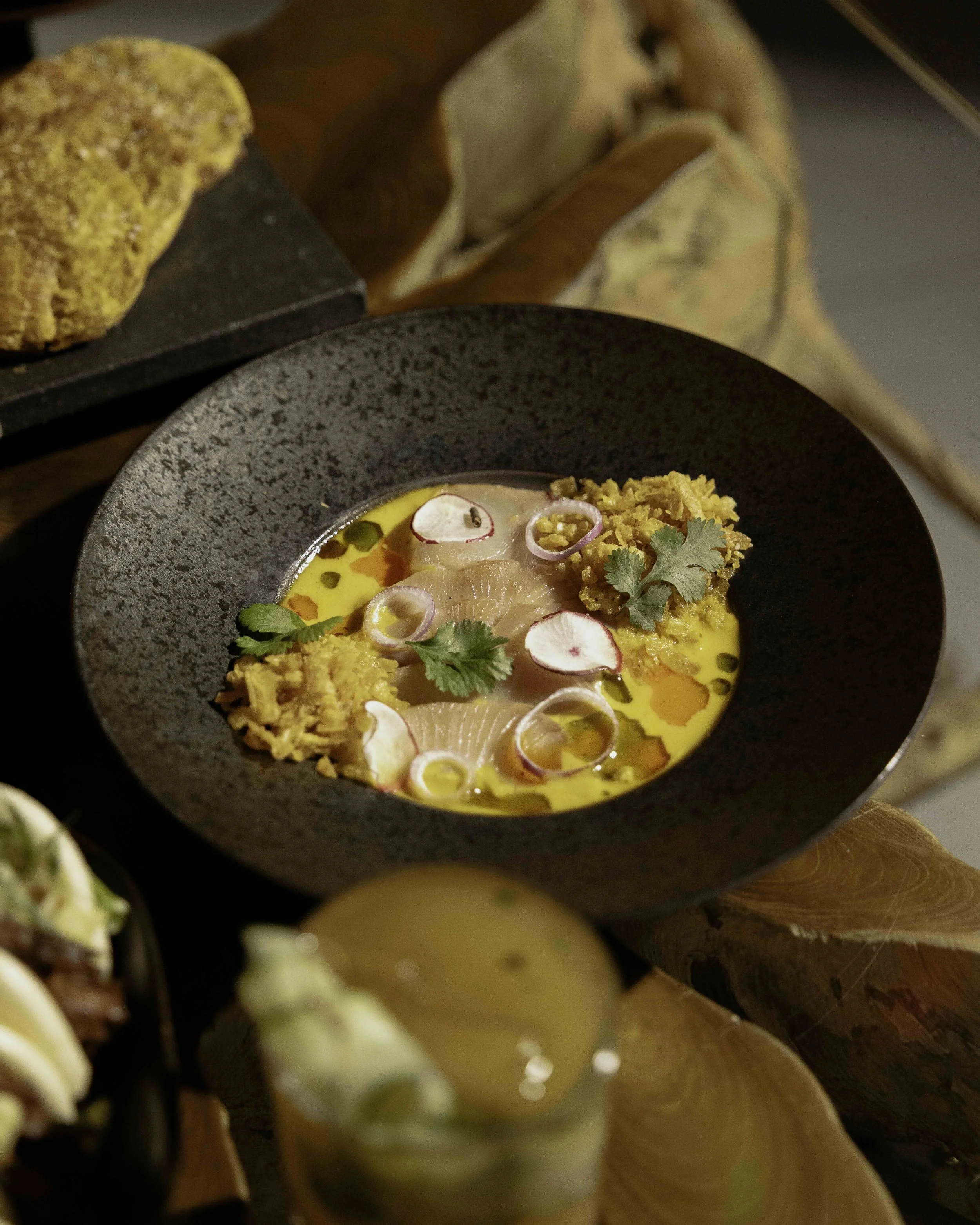A black bowl containing yellowish ceviche topped with radish slices, cilantro, and crispy tostadas, with a serving of bread and a drink in the background.