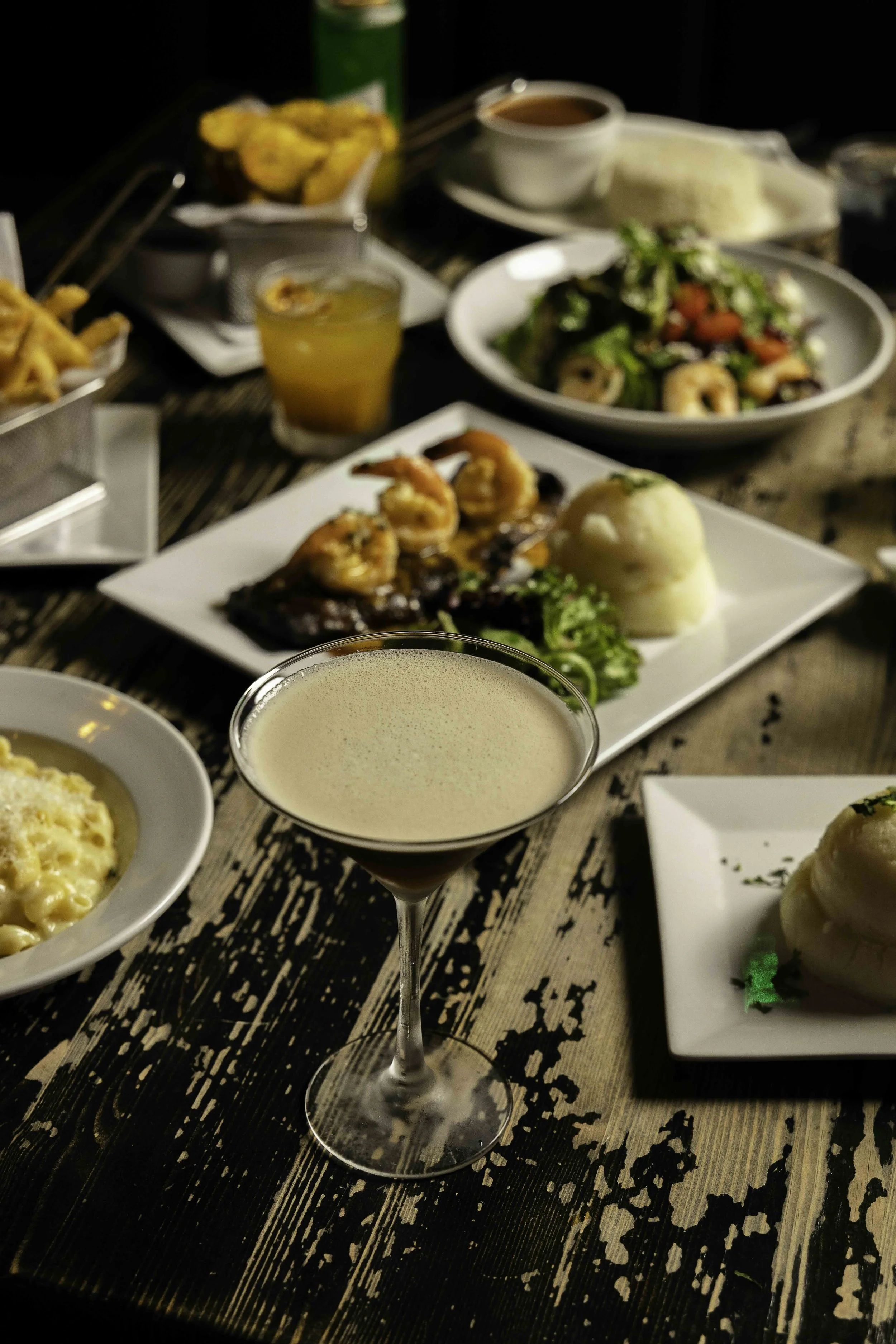 A table set with various dishes including a baked shrimp dish, a salad, mashed potatoes, and a creamy cocktail in a martini glass.