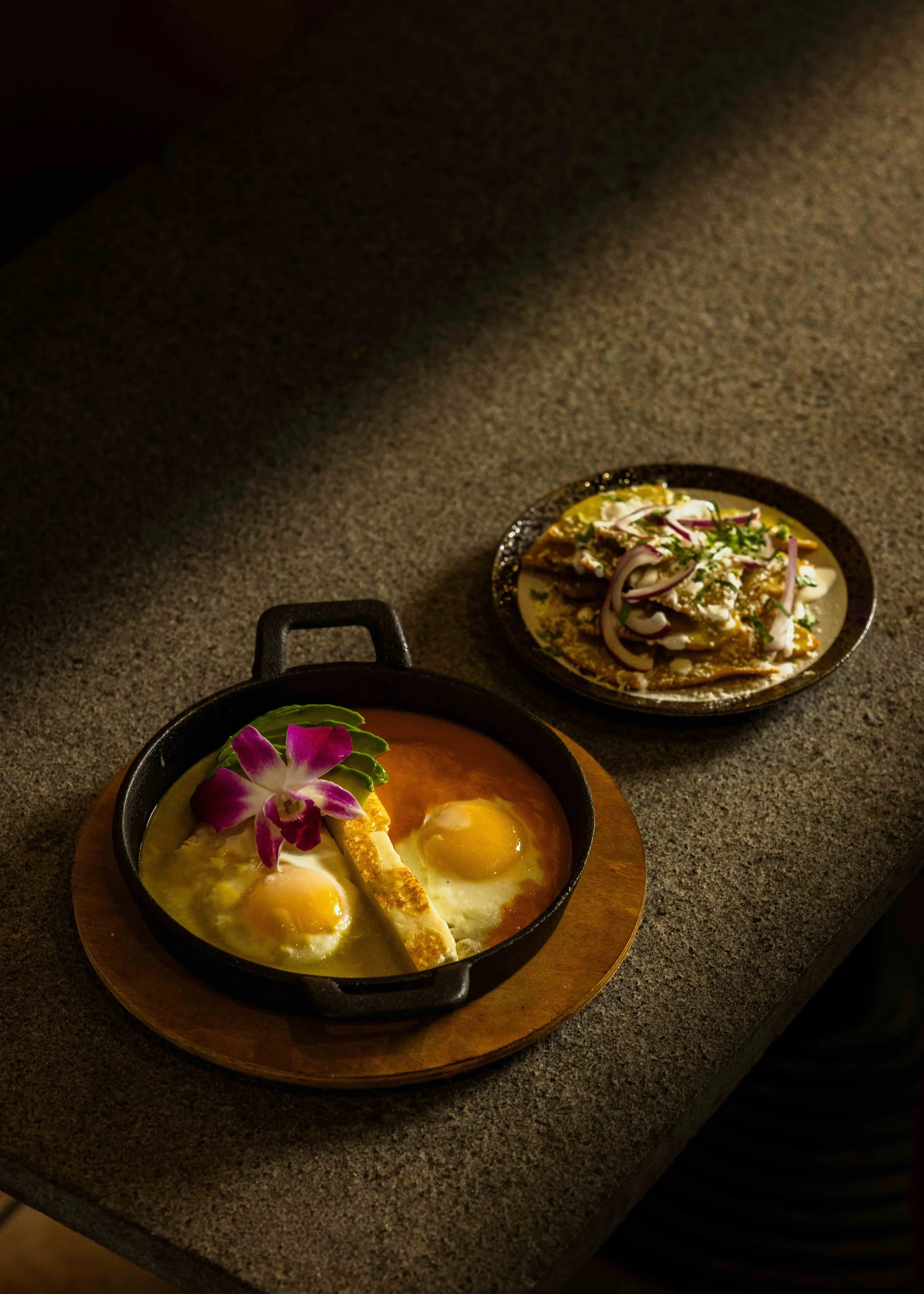 A black skillet with two sunny side up eggs, garnished with purple orchid and green leaf, placed on a wooden serving board. In the background, a plate with a dish topped with onions, herbs, and sauce, is partially visible on a speckled gray counterto