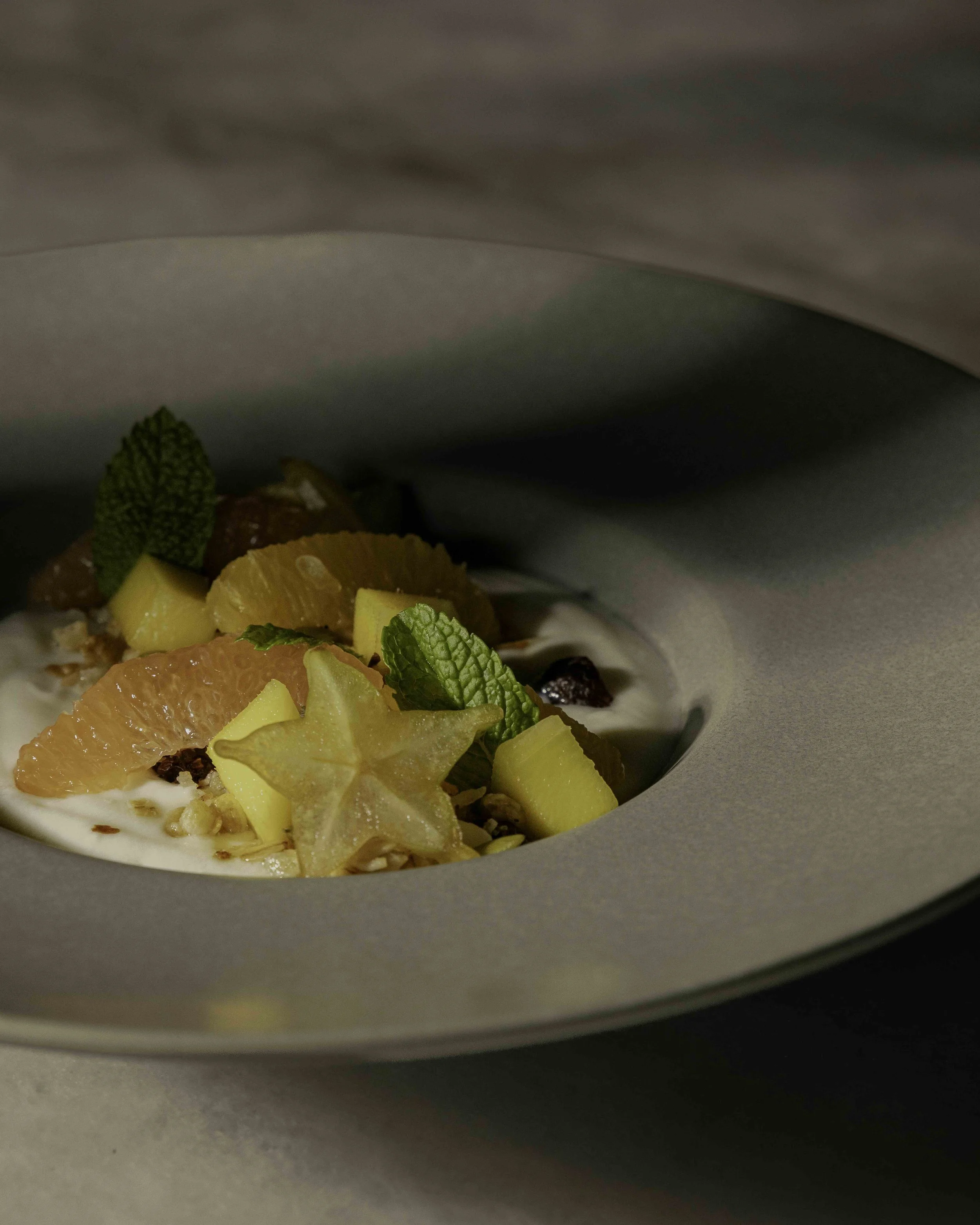 A dessert with fresh tropical fruits, mint leaves, and yogurt or cream in a gray bowl, partially covered by a gray cloth.