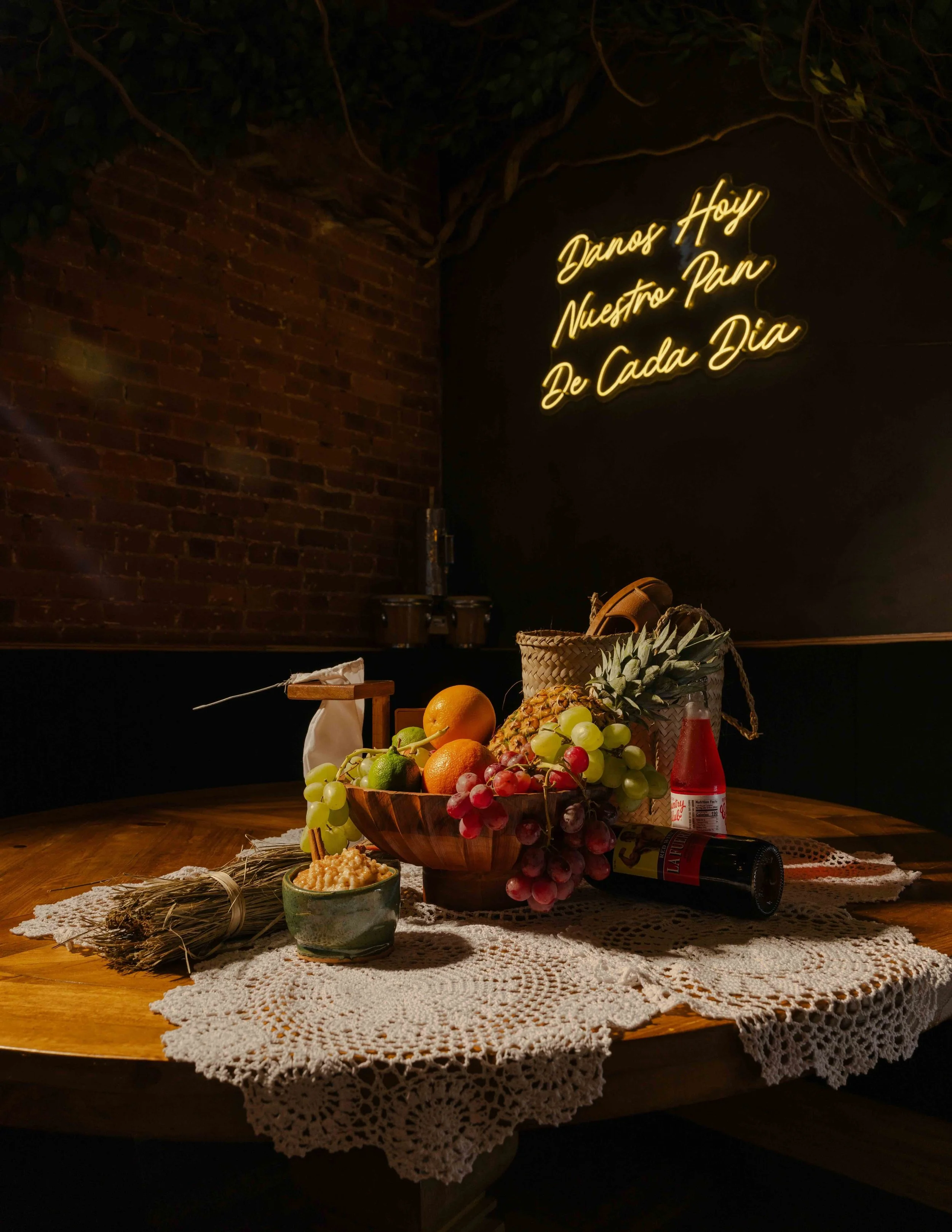 Neon sign reading 'Danos Hoy Nuestro Pan De Cada Dia' above a table with a bowl of assorted fruits, a bundle of dried herbs, a small container of cheese, a basket, and various bottles, set on a crocheted tablecloth.