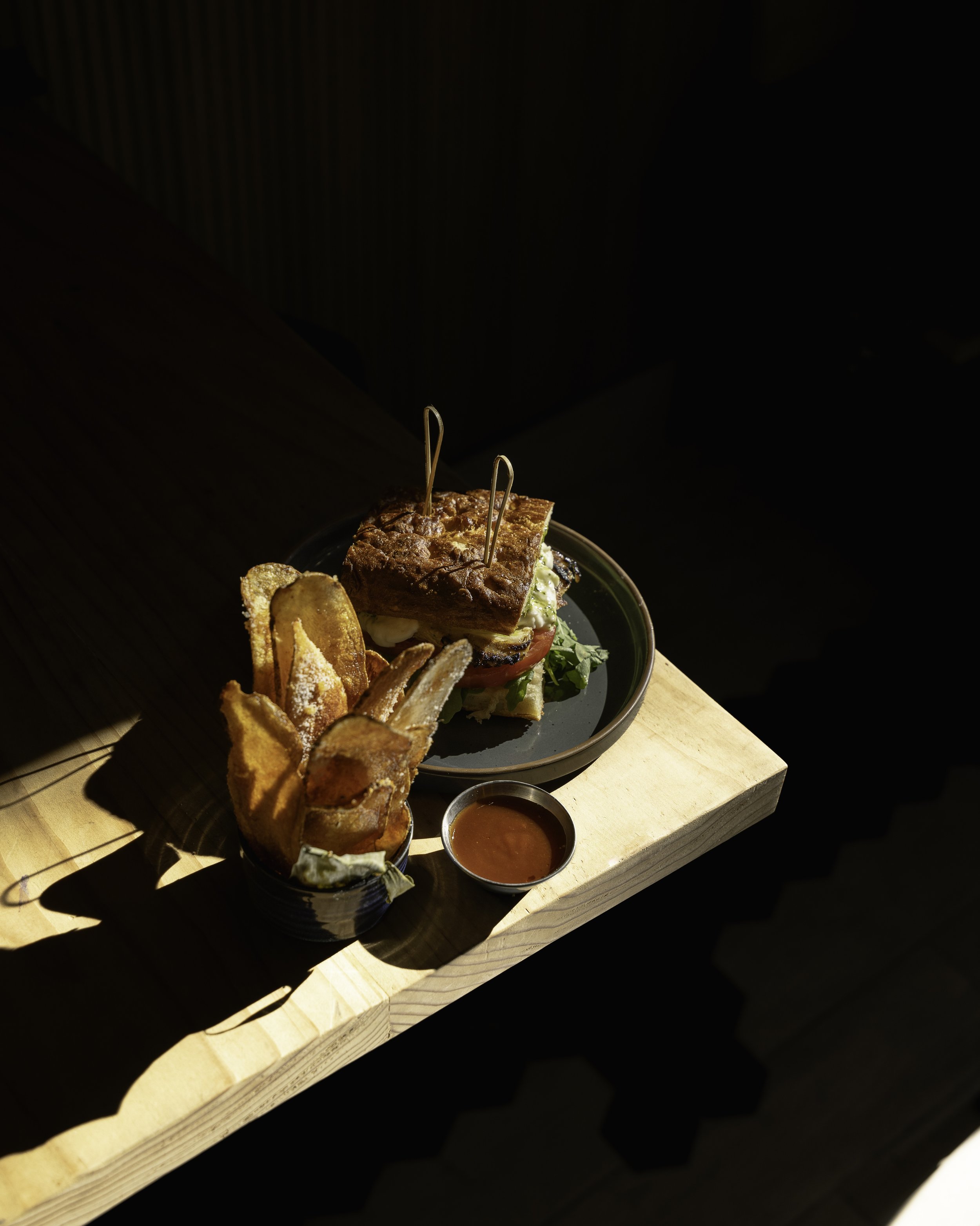 A sandwich with fried chicken, lettuce, tomato, and cheese served with potato chips and dipping sauce on a black plate, with additional chips and sauce on a small black bowl and a cup on a wooden surface.