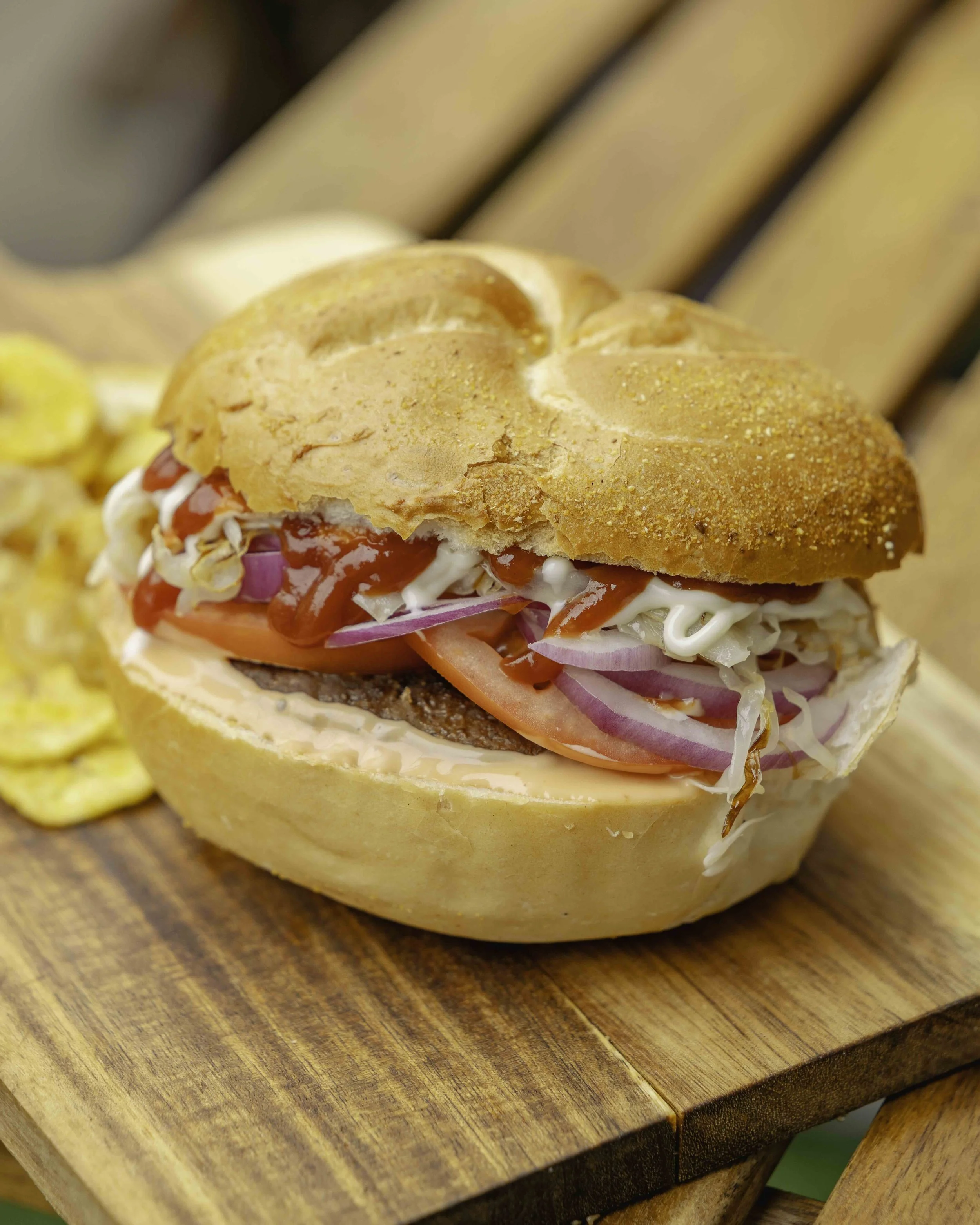 A cheeseburger with tomato, onion, and condiments on a wooden surface with potato chips to the side.