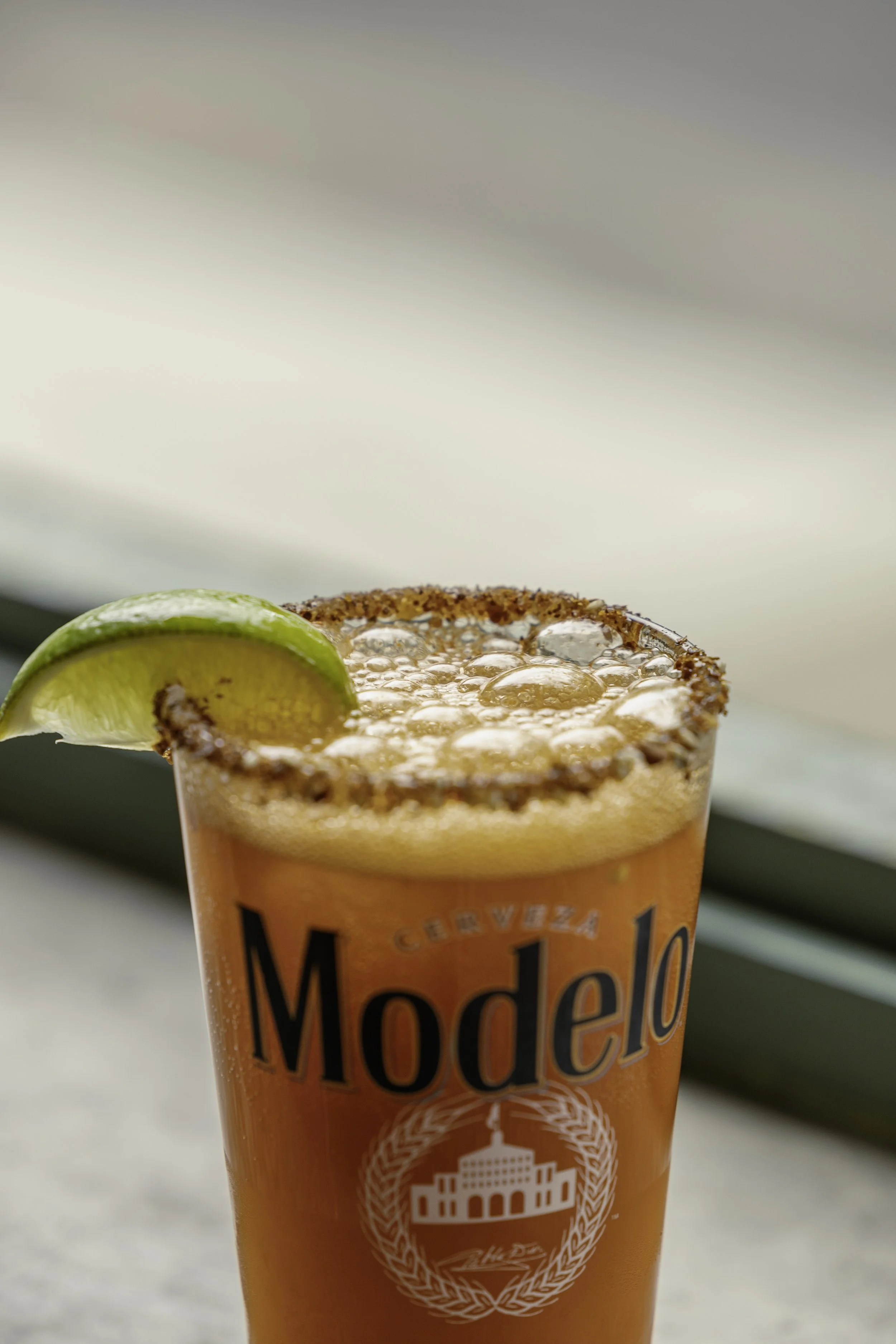 Close-up of a Margarita cocktail in a glass with a salted rim and a lime wedge garnish, with the Modelo logo on the glass.