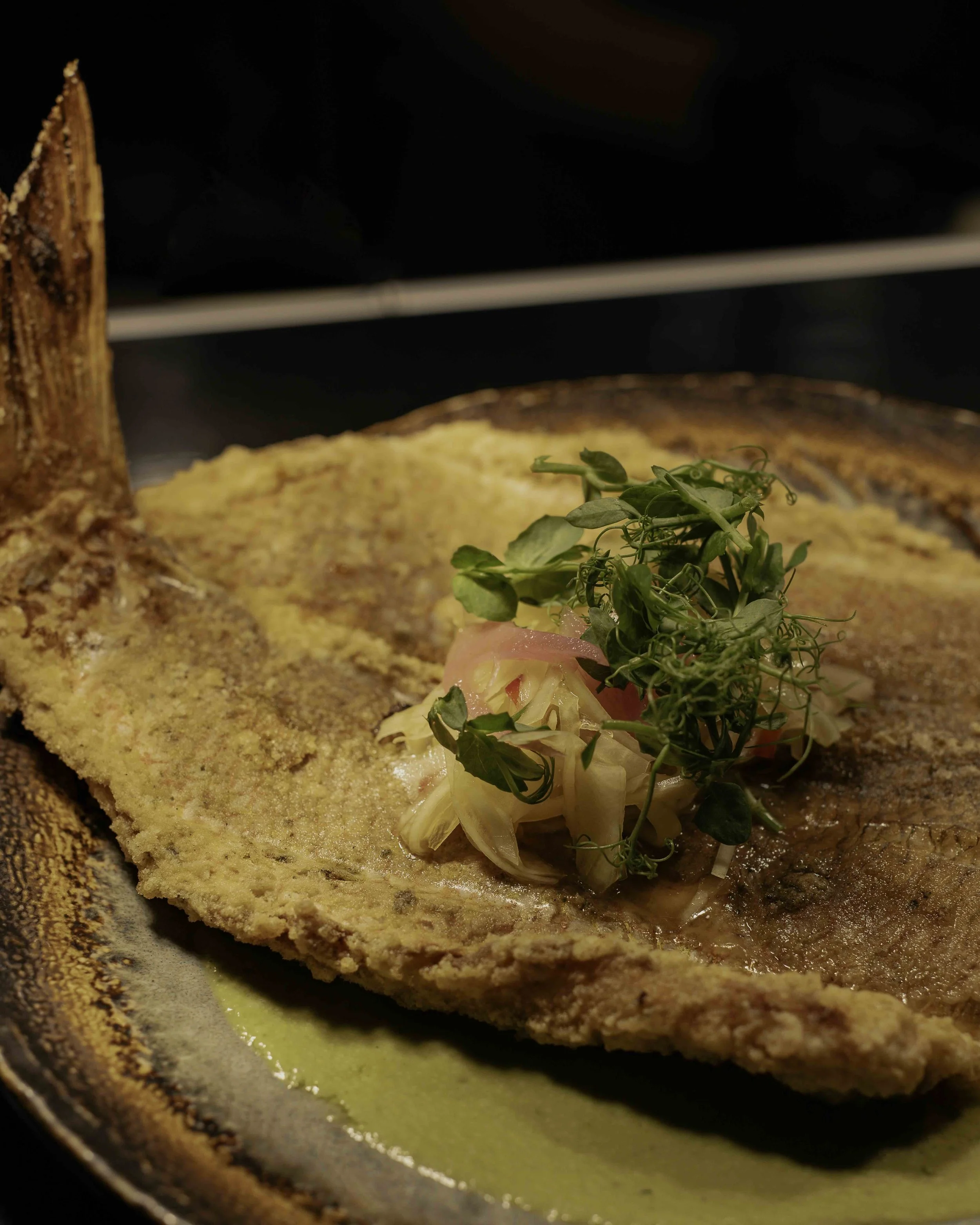 Close-up of a cooked fish fillet topped with chopped onions and fresh herbs, served on a rustic plate with a drizzle of yellow sauce.