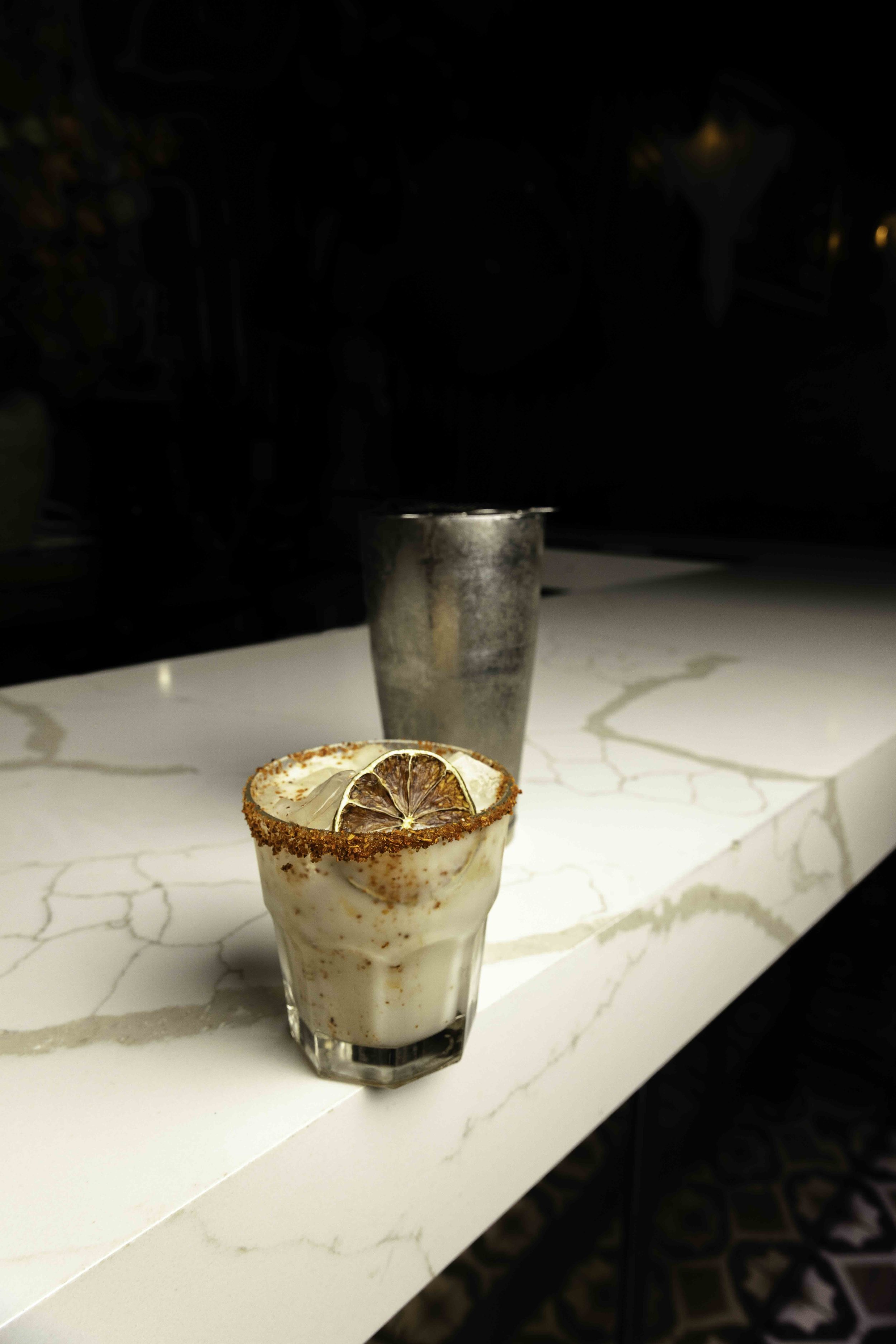 A creamy cocktail drink garnished with dried orange slices and a chili powder rim, placed on a white marble countertop with a metallic cup in the background.