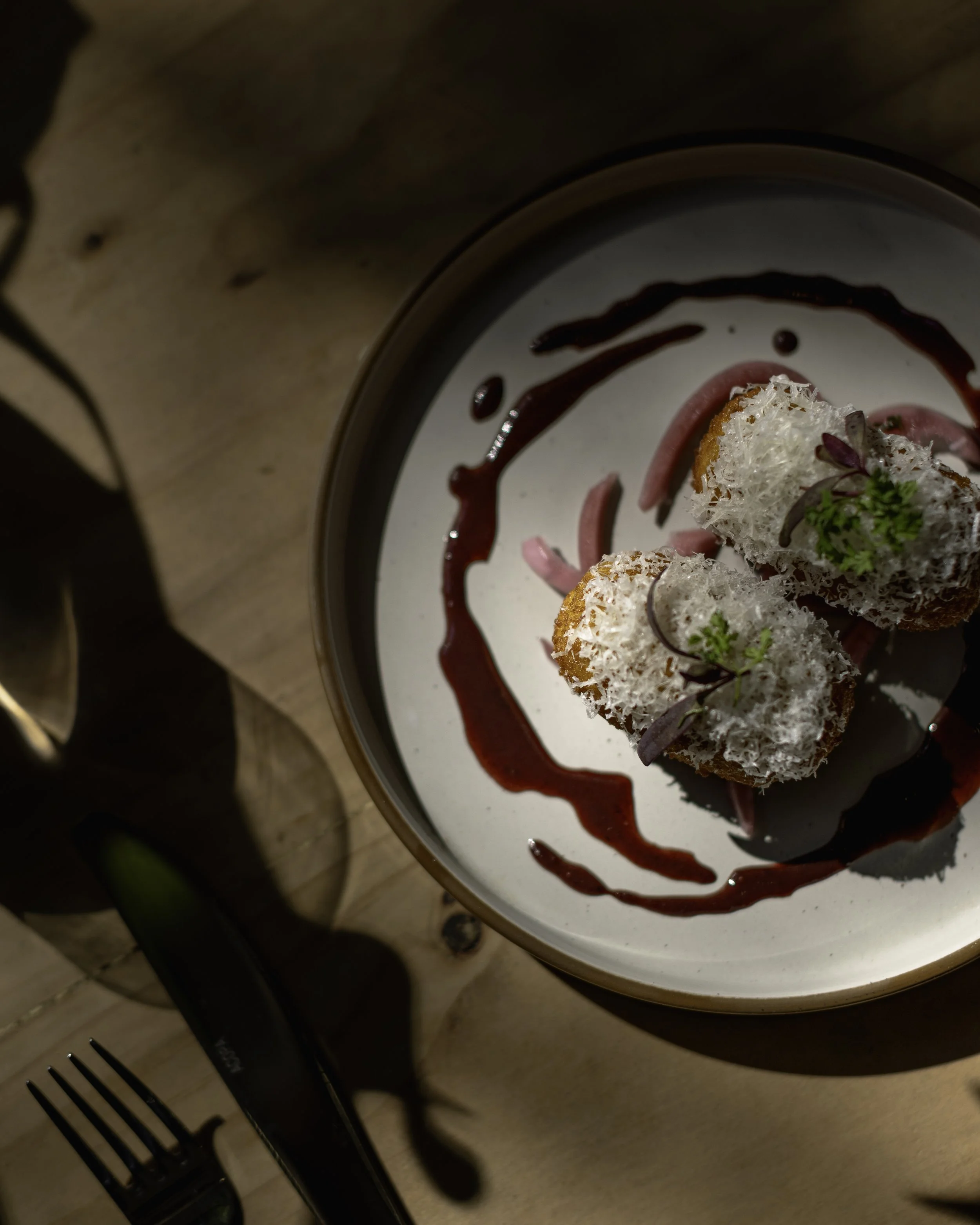 A plate with two croquettes garnished with herbs and a drizzle of red sauce, served on a white surface with decorative sauce art.