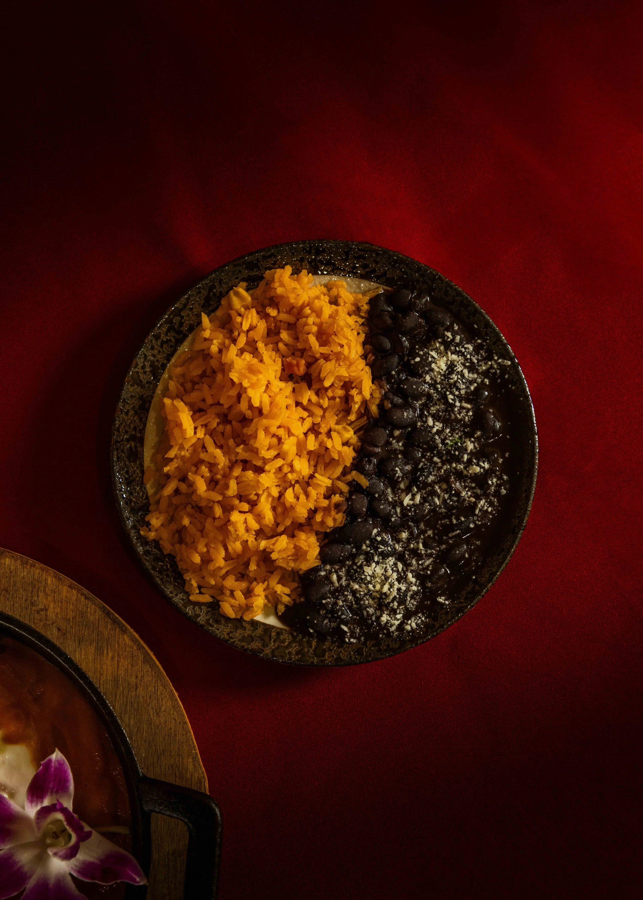 Bowl of Mexican food with yellow rice, black beans, and grated cheese on top, placed on a red tablecloth.
