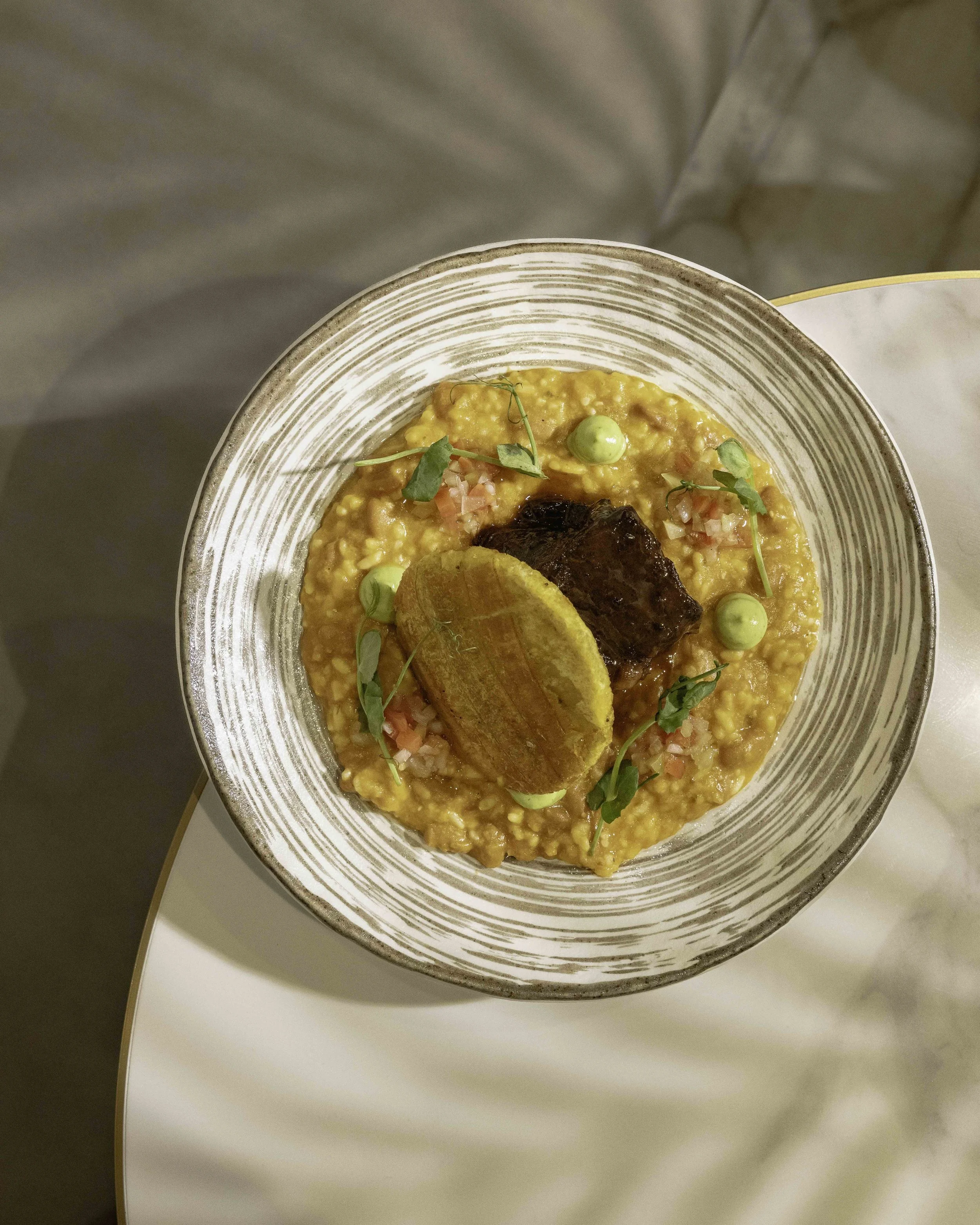 A bowl of yellow risotto garnished with green pea sprouts, diced vegetables, a black piece of meat, and toasted bread slices.