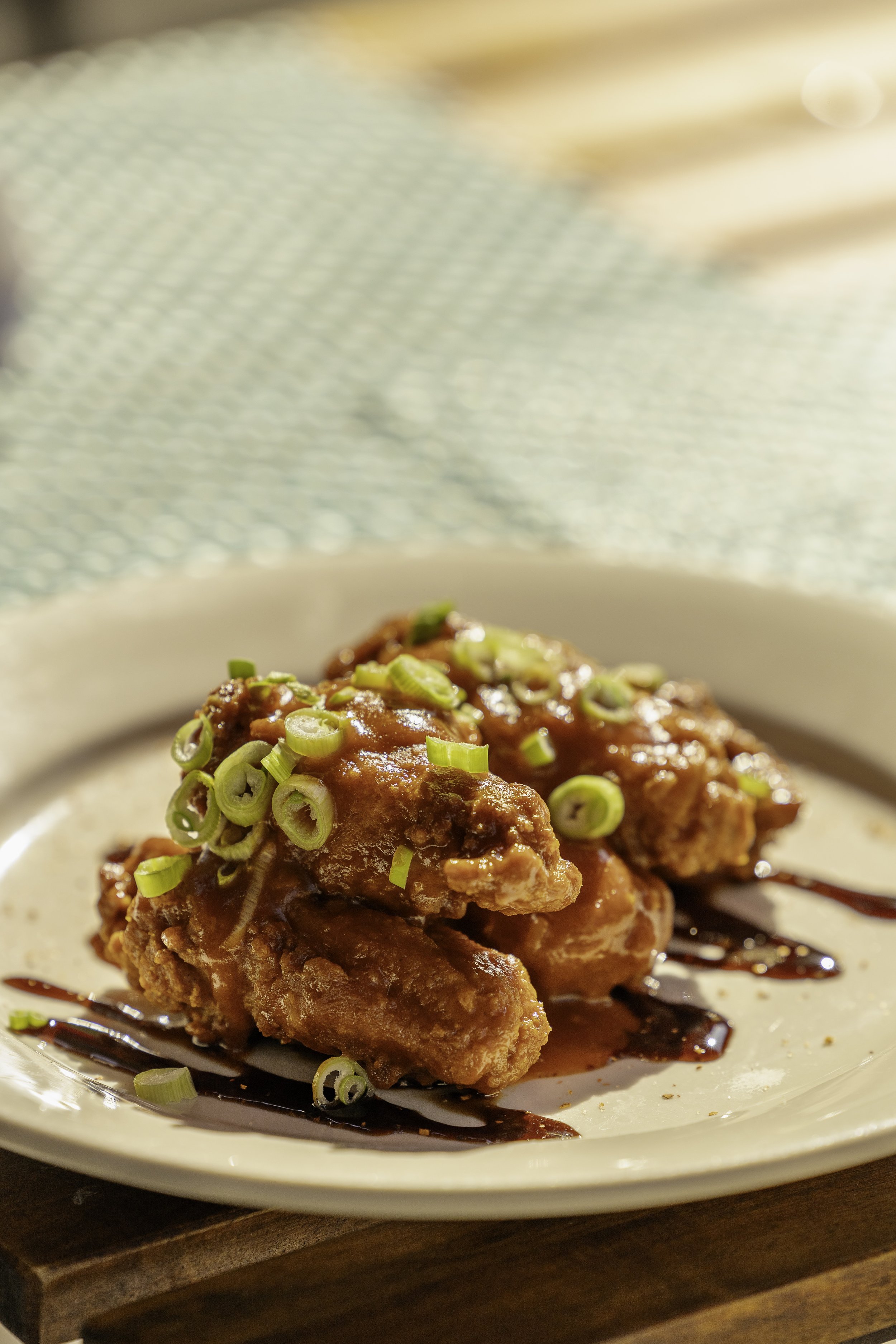 Plate of fried chicken topped with green onions, drizzled with dark sauce, on a white plate with a wood coaster.