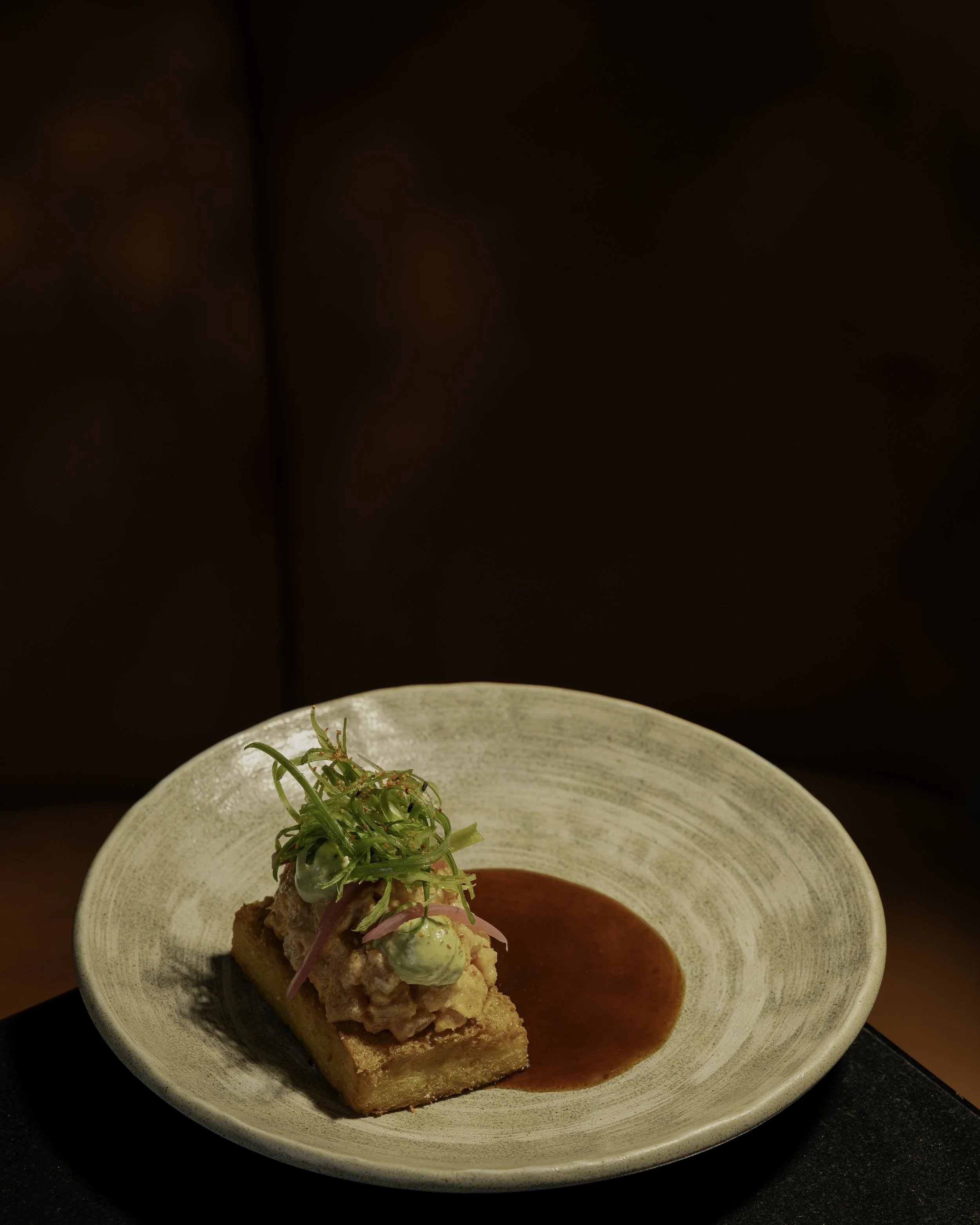 Close-up of gourmet appetizer with a toasted bread base topped with a creamy seafood mixture, garnished with microgreens, served with a side of red sauce on an off-white ceramic plate.