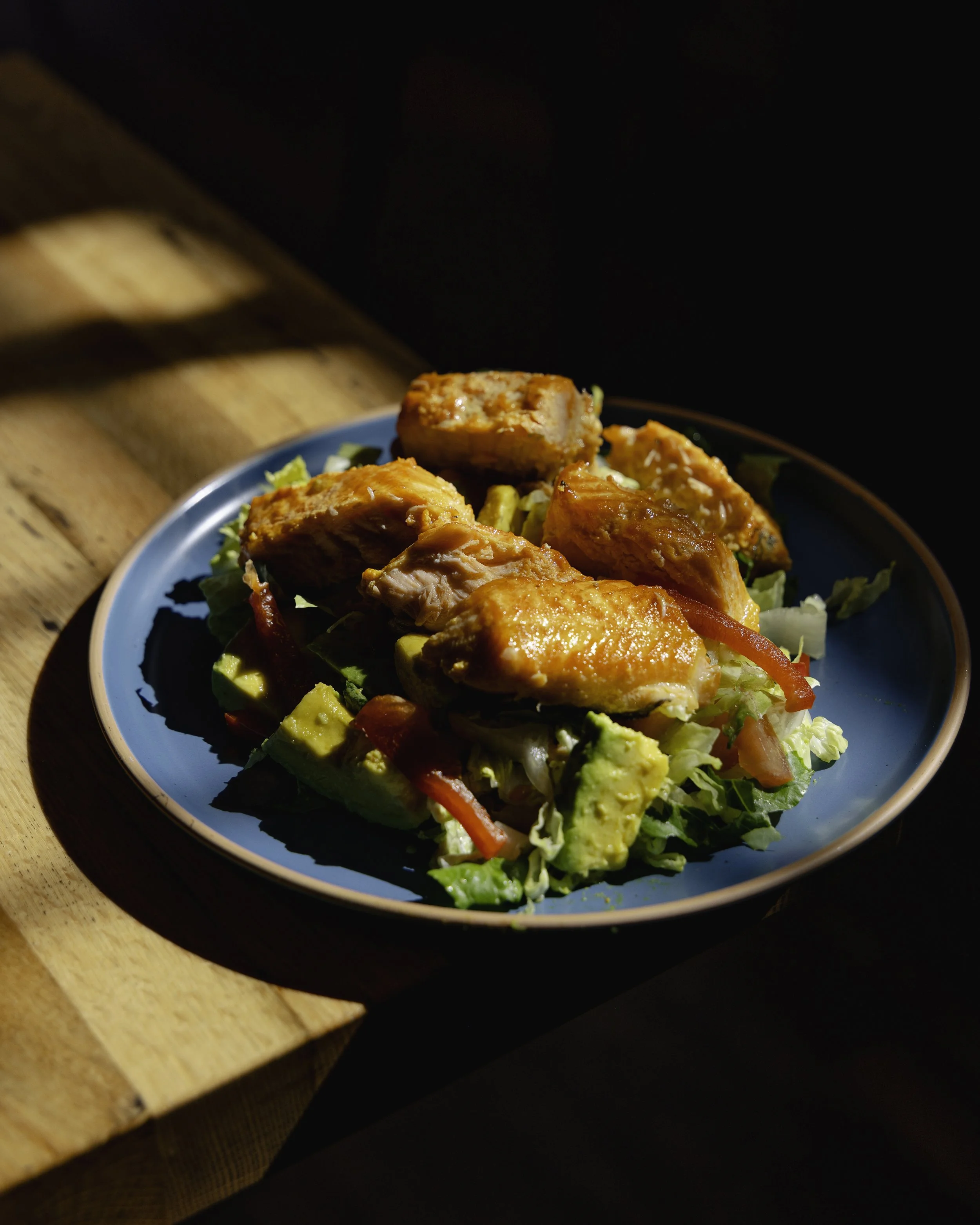 A blue plate with grilled chicken pieces atop a salad with avocado, lettuce, and red bell peppers, placed on a wooden surface with sunlight casting shadows.