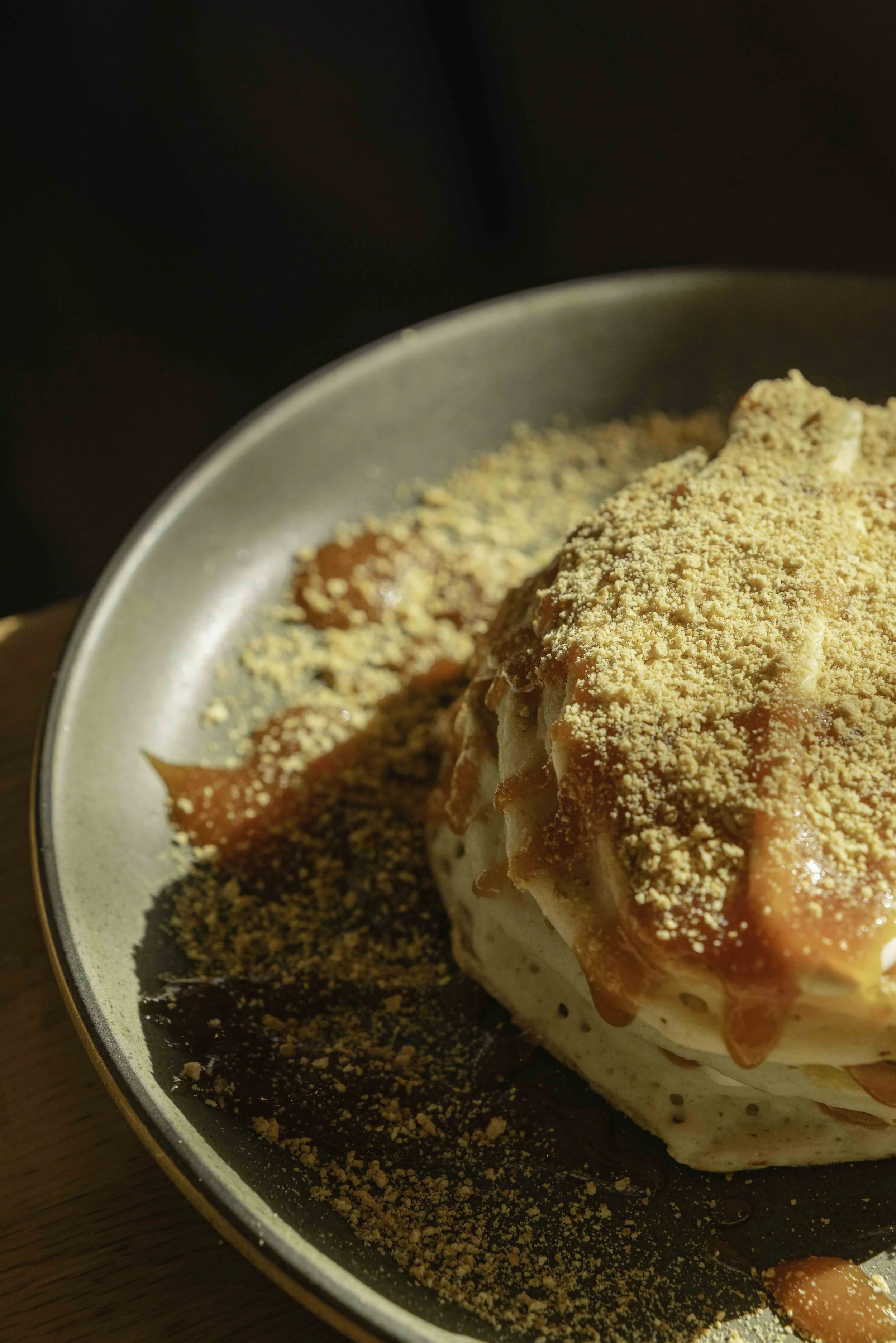 Close-up of a dessert topped with crushed topping and drizzled with caramel sauce on a dark plate.