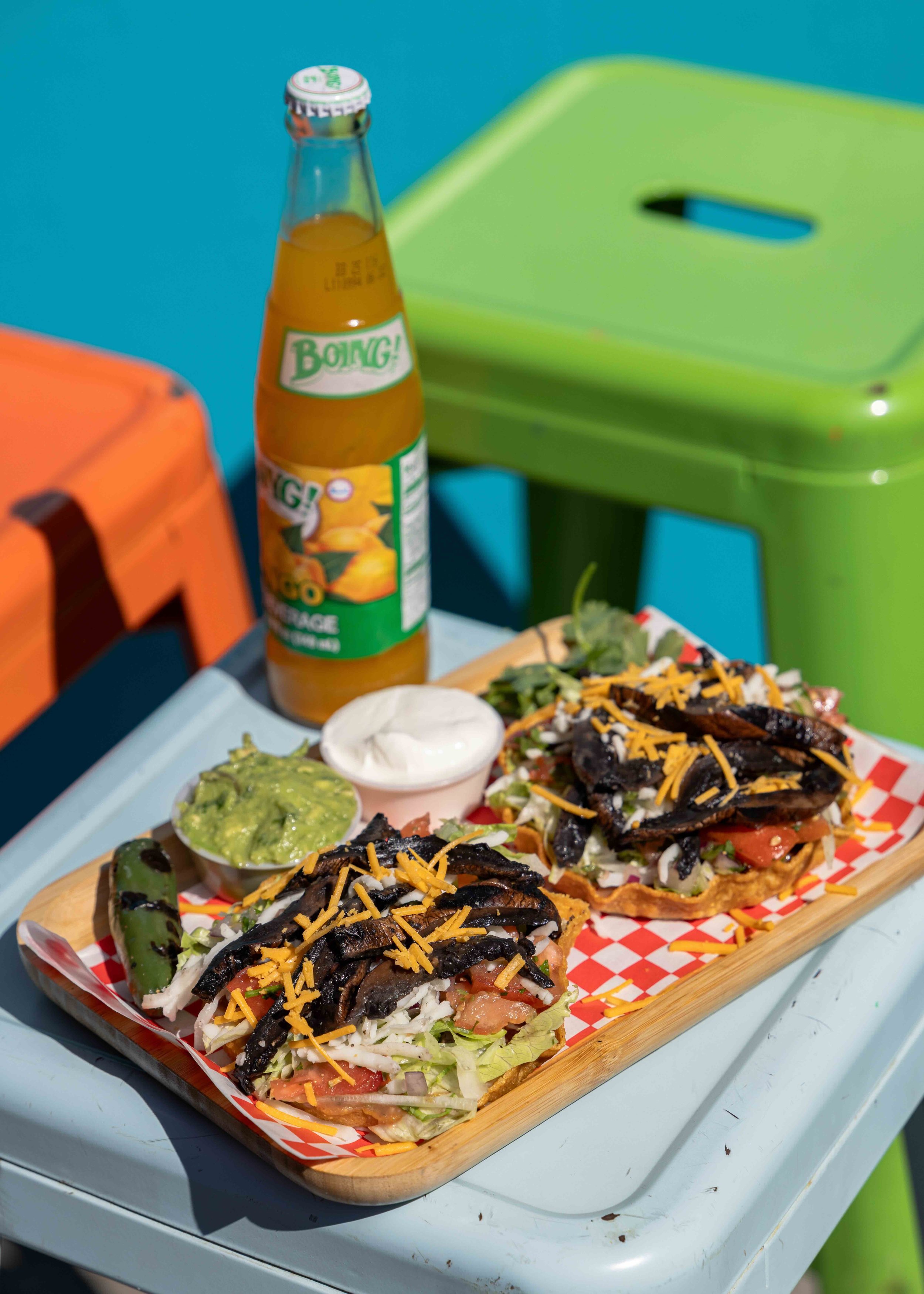 Two tacos with grilled vegetables, shredded cheese, lettuce, chopped tomatoes, and sour cream, served on a wooden platter with guacamole, a jalapeño, a bottle of mango-flavored drink, and a container of sour cream, on an outdoor table.