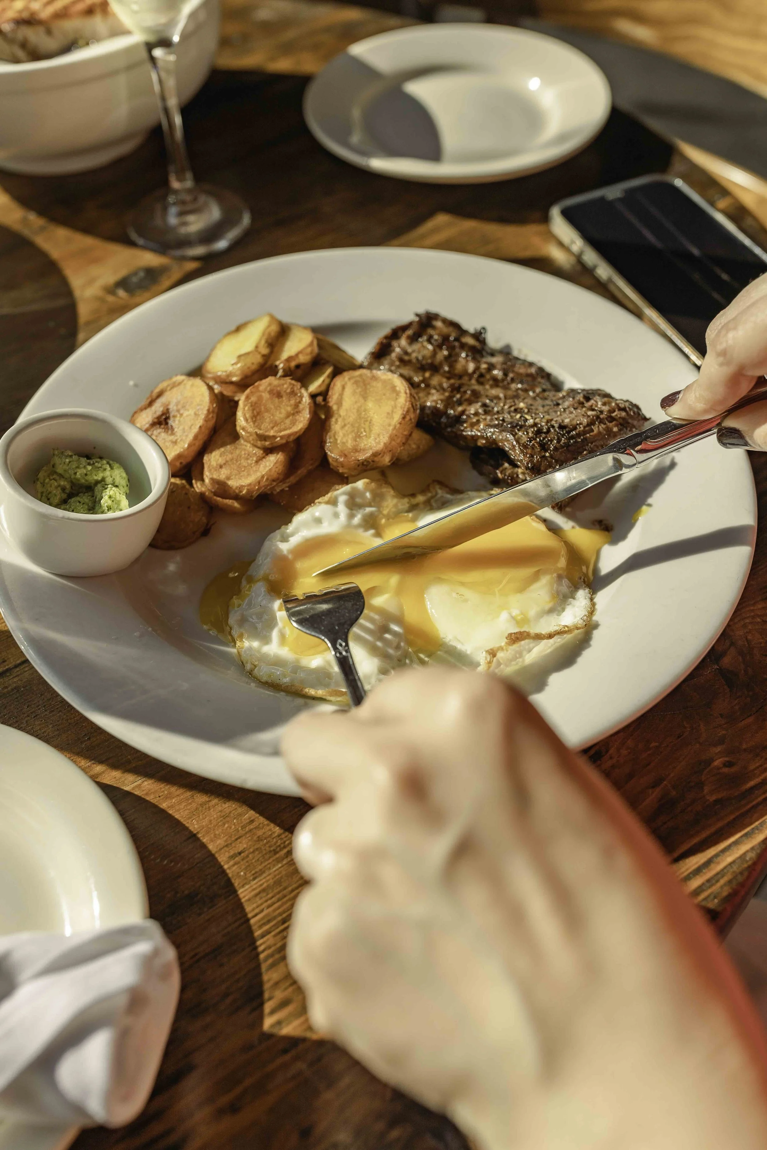 A breakfast plate with fried eggs, potato wedges, grilled steak, and a small serving of guacamole, set on a wooden table with eating utensils and a glass of wine.