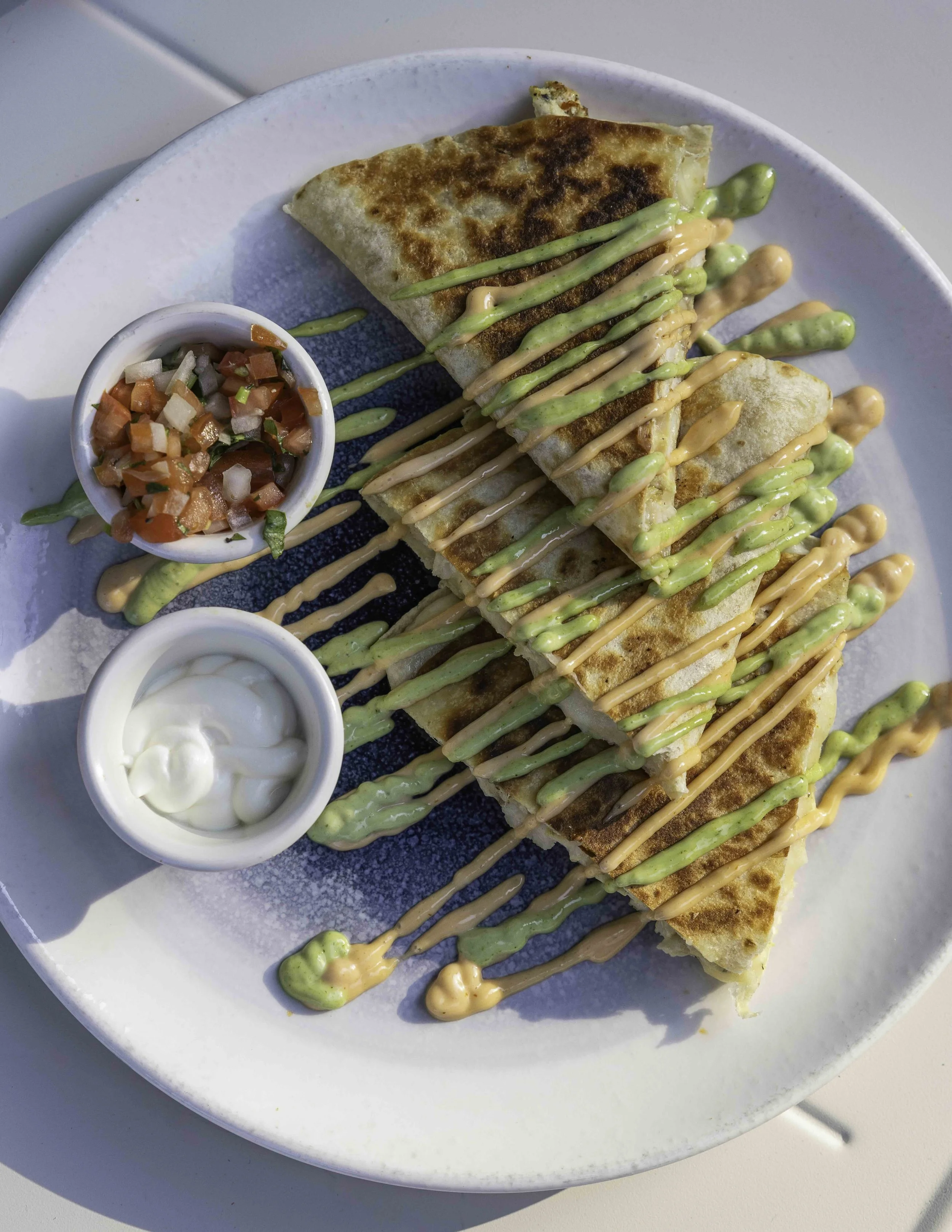 Plate of quesadillas drizzled with green and orange sauces, served with small bowls of salsa and sour cream.