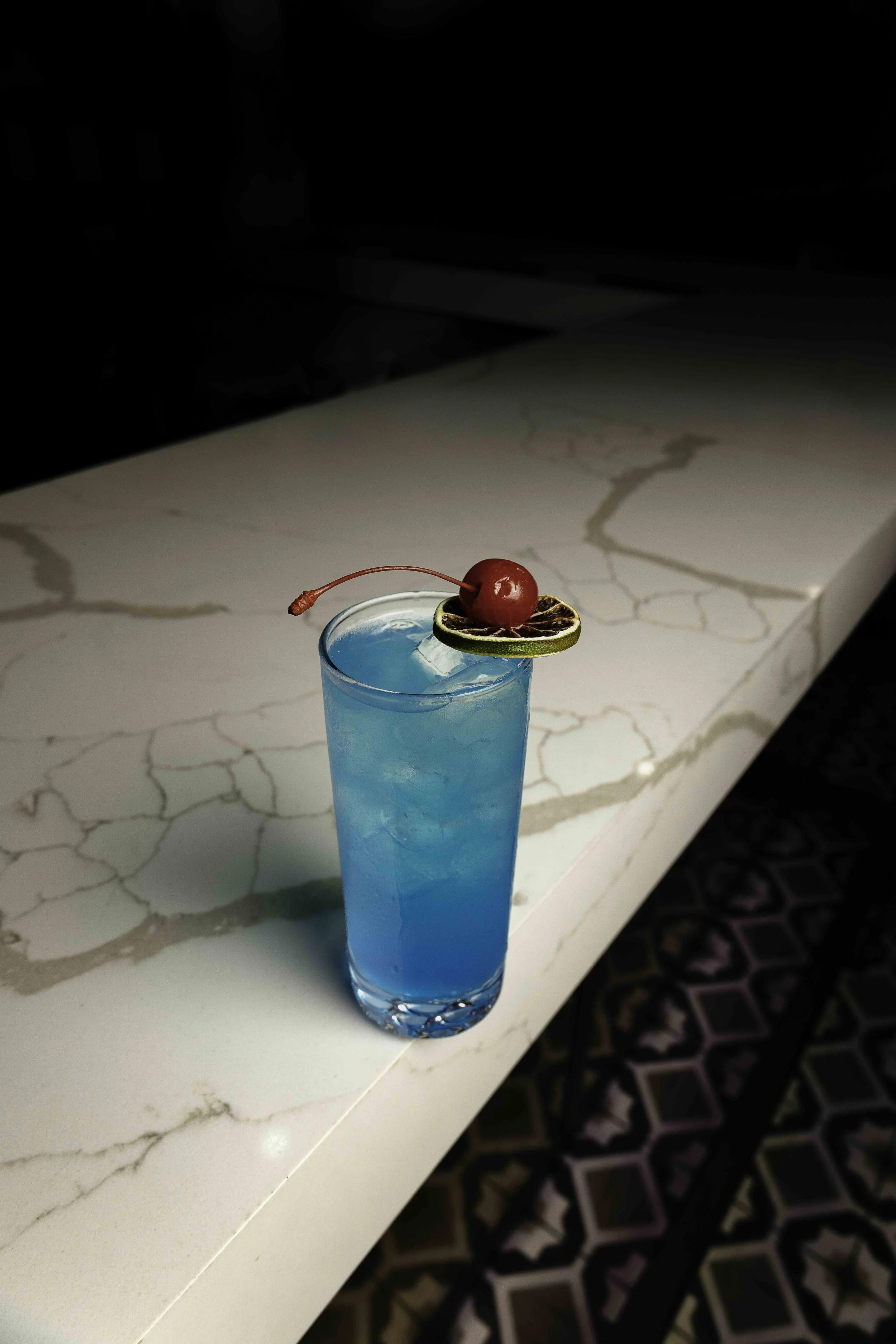 Blue cocktail in a tall glass garnished with a cherry and a slice of lime, sitting on a white marble countertop with a dark background and colorful patterned carpet.