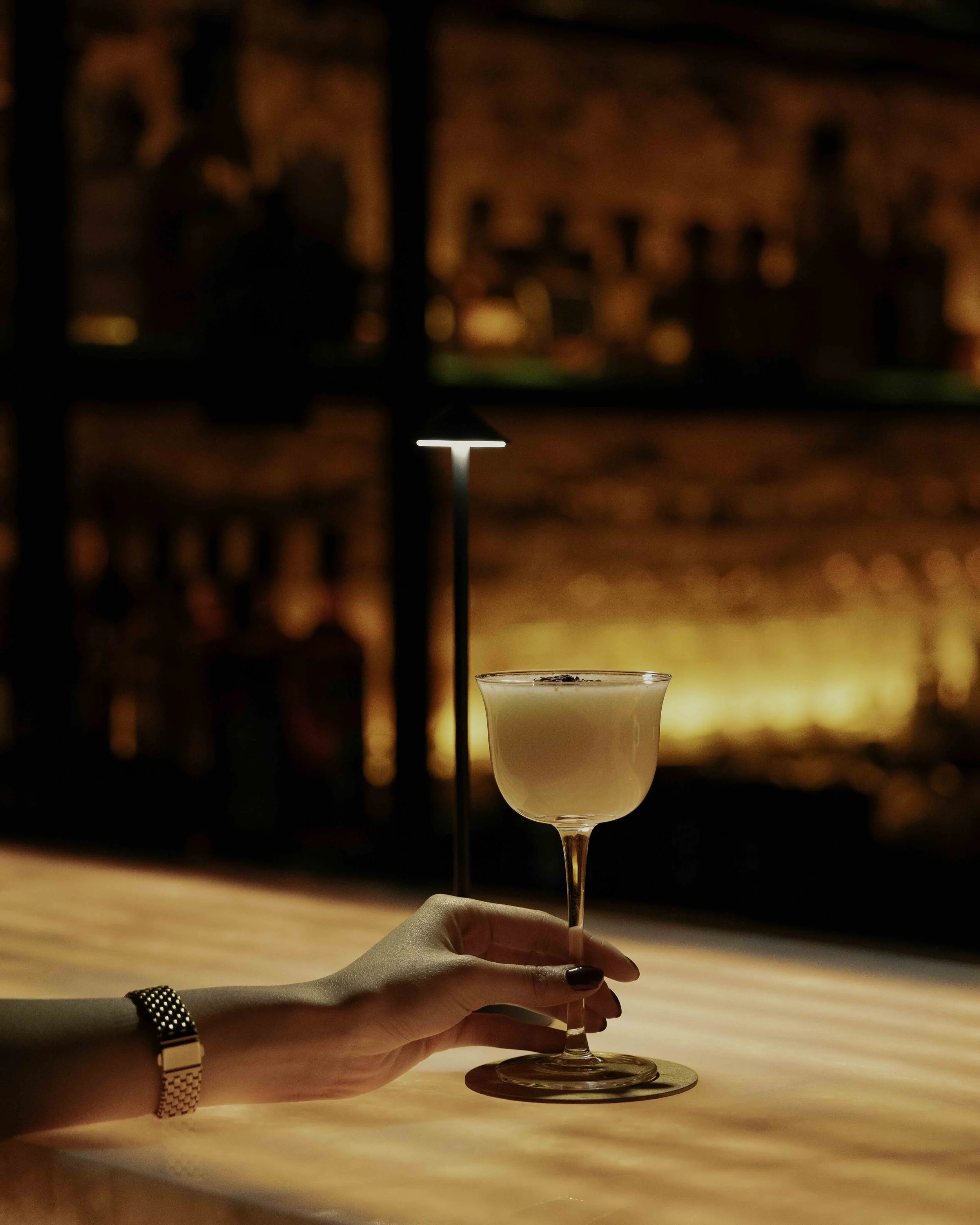 A person's hand holding a cocktail glass with a creamy drink, in a dimly lit bar or lounge with warm lighting and a window showing a blurred cityscape at night.