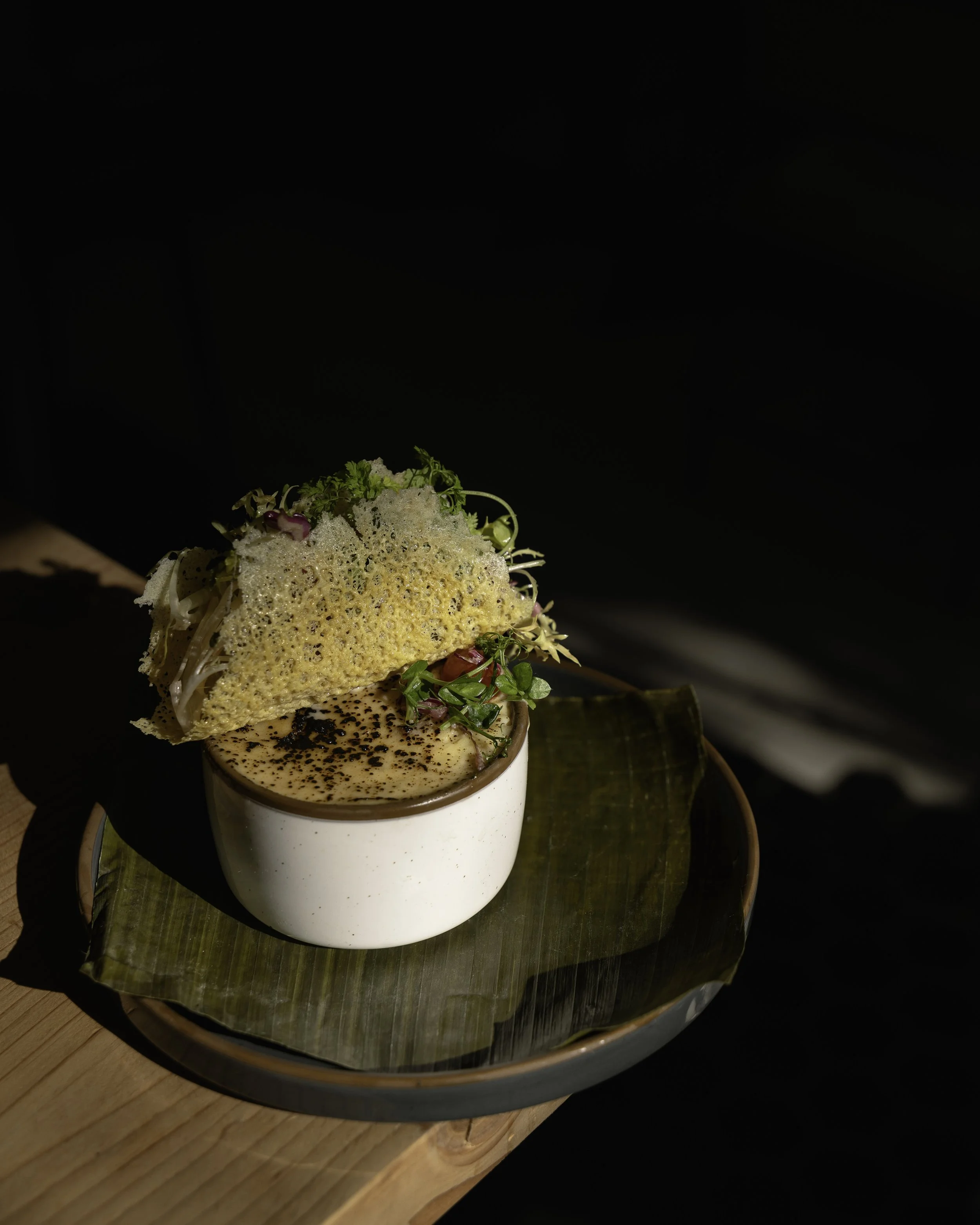 A food dish featuring a cup of coffee garnished with cocoa powder, served with a crispy, bubbly bread topped with microgreens and edible flowers on a banana leaf, on a wooden table.