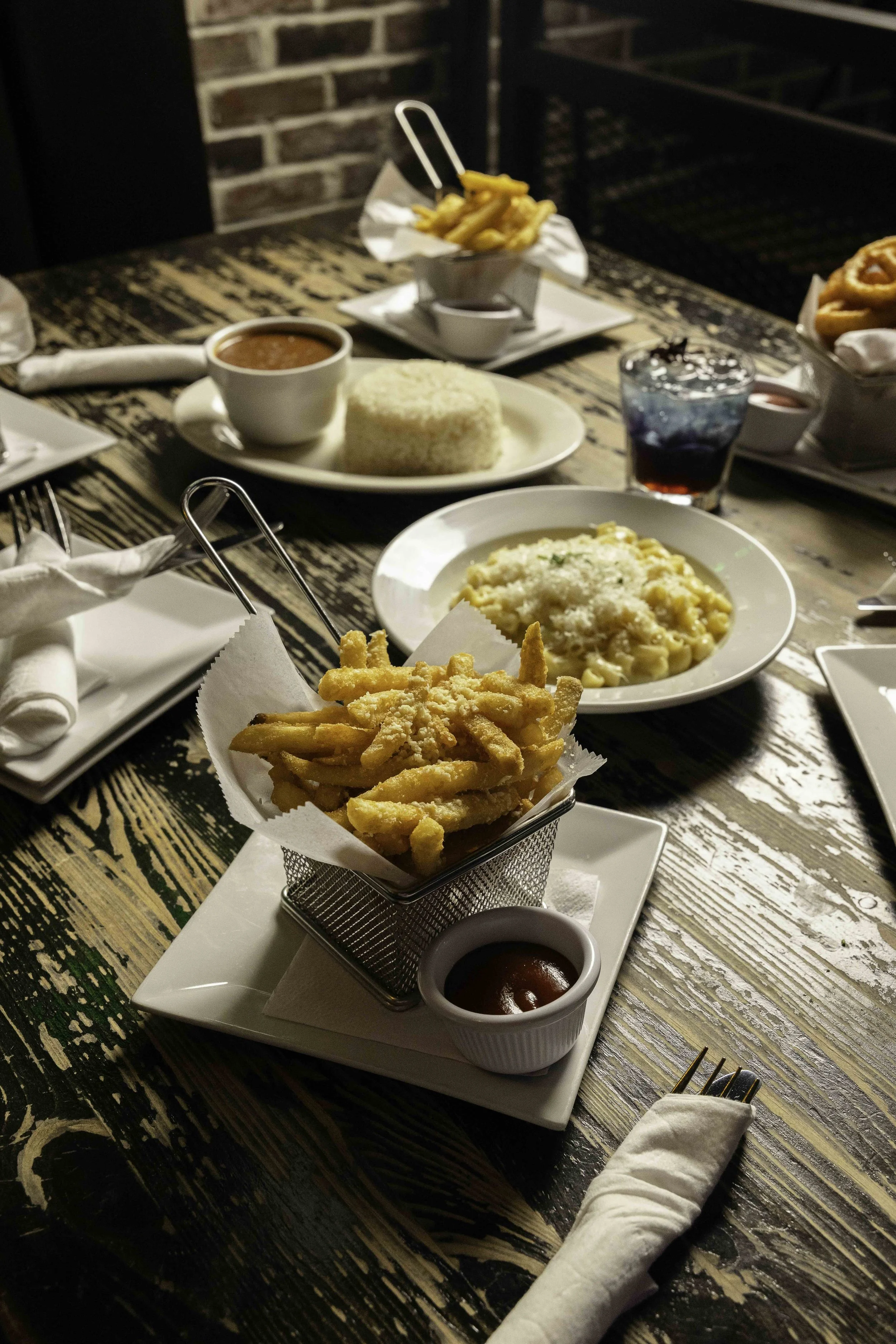 Fried French fries with ketchup, macaroni and cheese, bowl of tomato soup with rice, cheeseburger pasta, and a blue cocktail on a rustic wooden table at a restaurant.